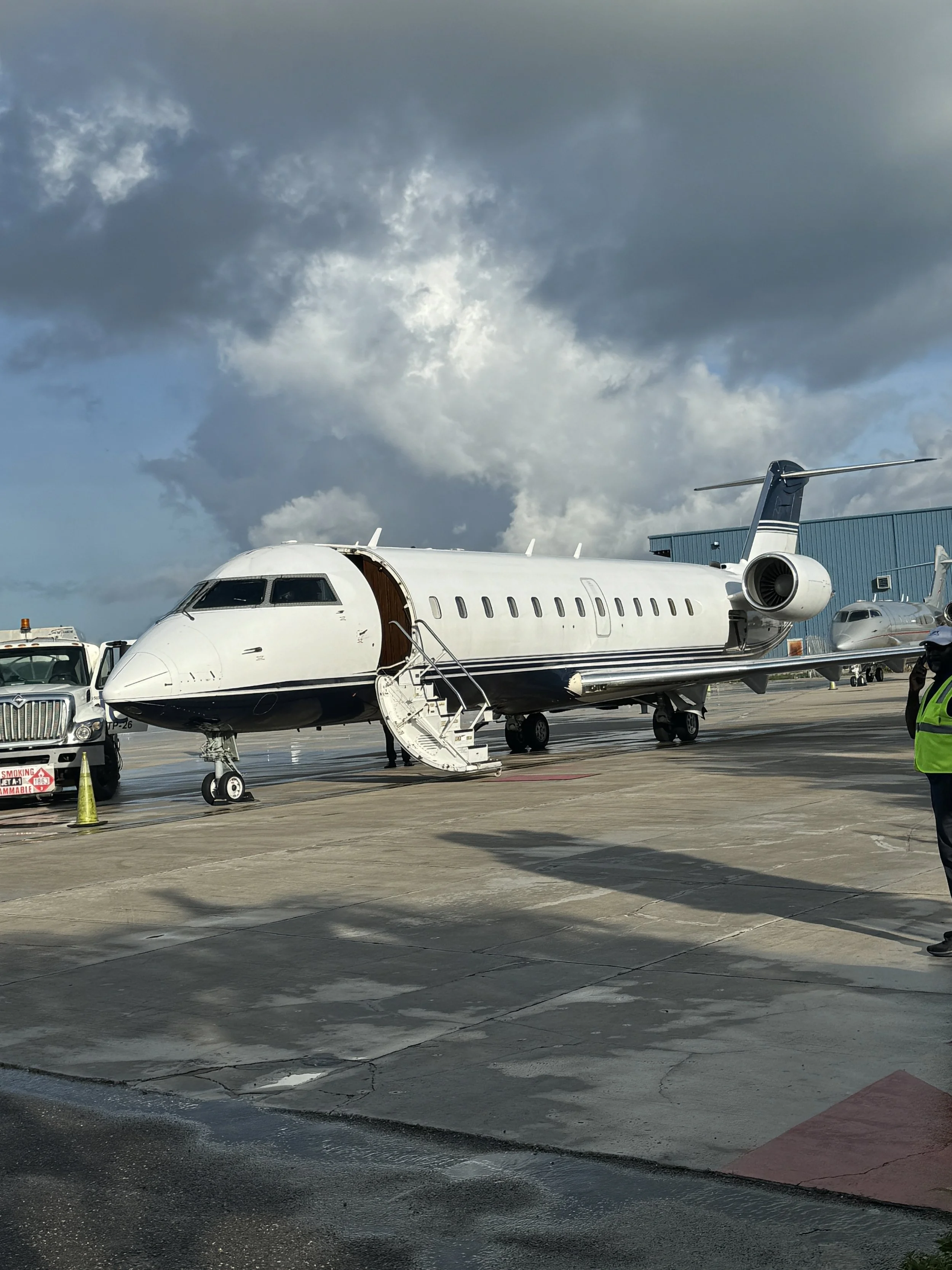 A private jet parked on an airport tarmac with its entrance stairs down. A white truck is nearby, and other aircraft are visible in the background under a cloudy sky.