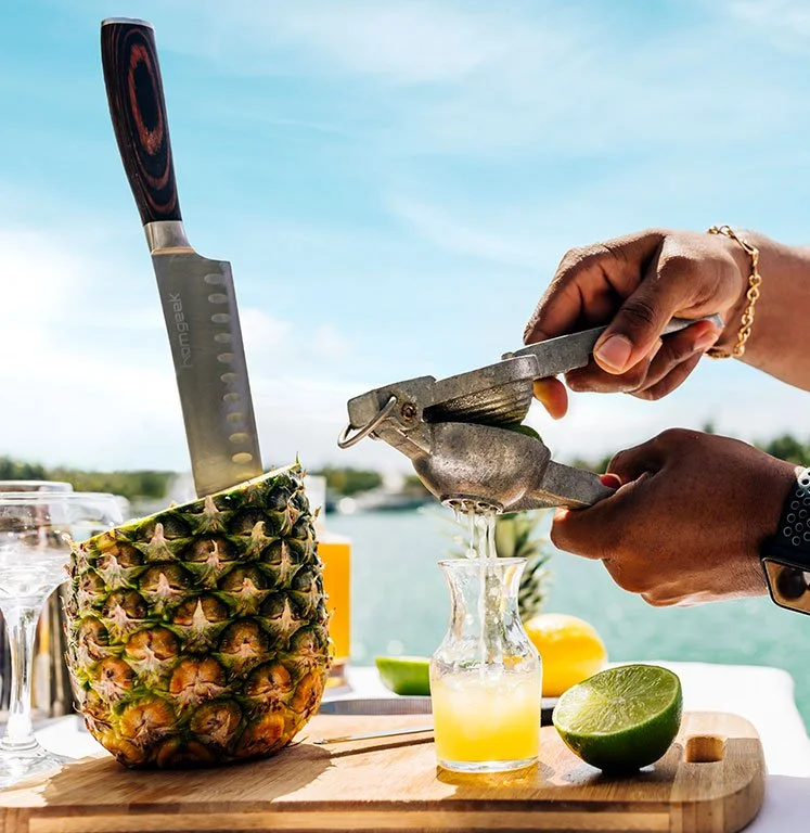 A person slicing a fresh pineapple into a glass jar with a citrus juicer, with cut lime and lemon in the background on a wooden surface near a body of water.