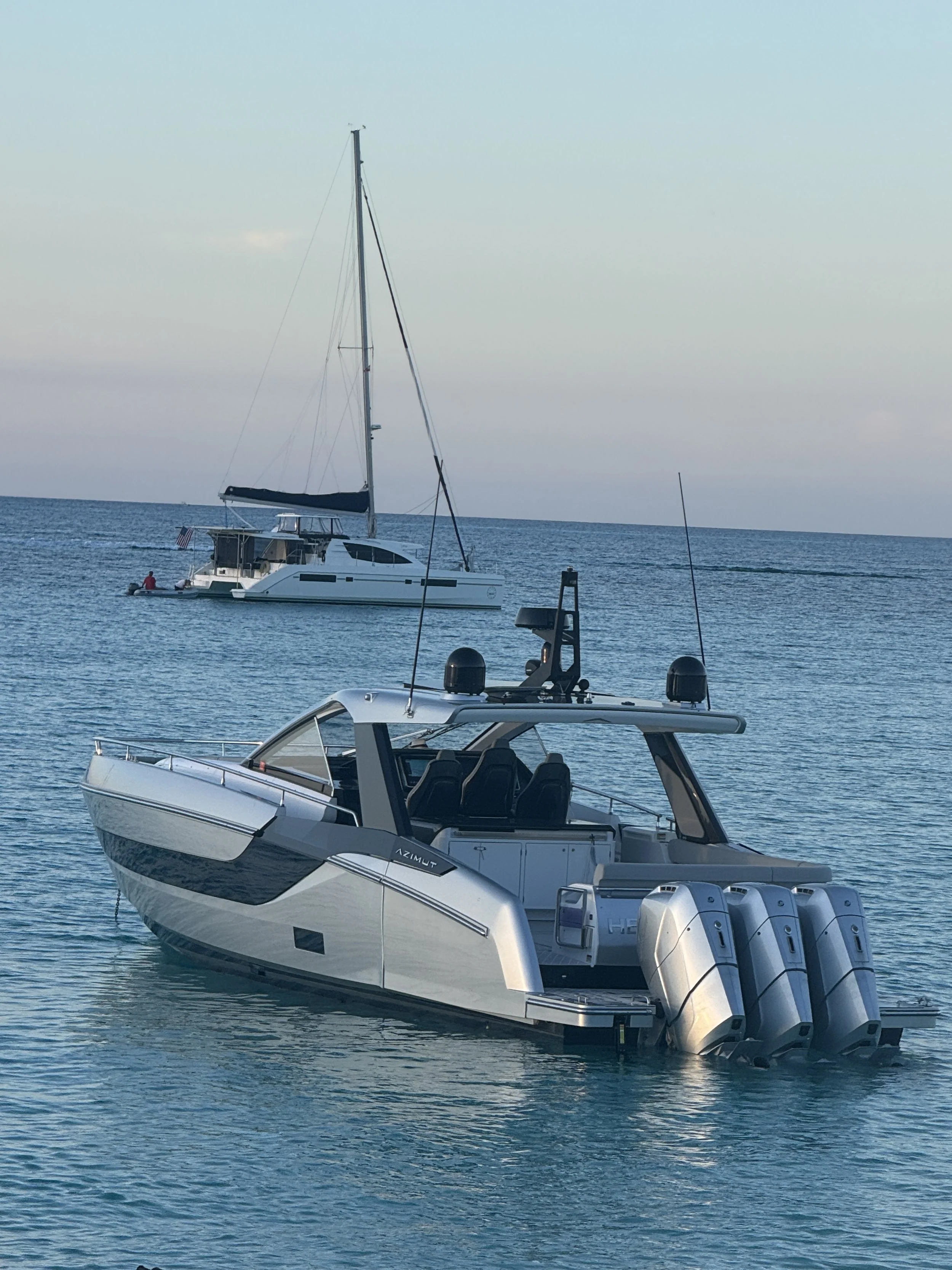 A modern white motor yacht with four outboard engines docked on calm water, with a sailboat in the background and a clear sky.