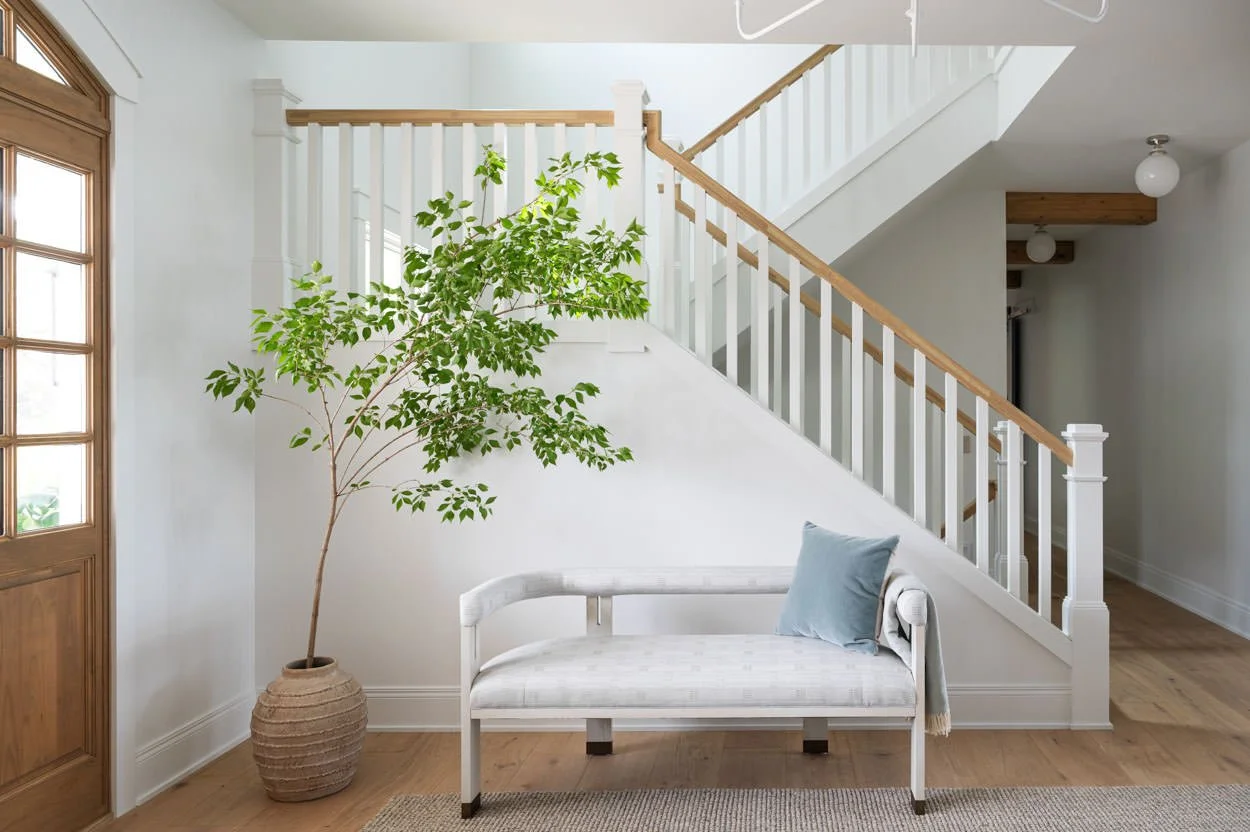 Interior foyer of a bright house with a white staircase, a potted plant, a white bench with a pillow, and wooden flooring. Full service interior design by KED Interiors