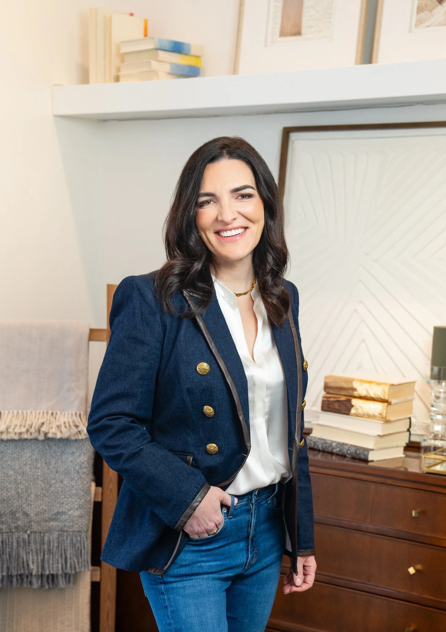 A woman with dark hair and a bright smile, wearing a navy blazer with gold buttons and blue jeans, standing indoors in front of a desk with stacks of books and a white textured wall.