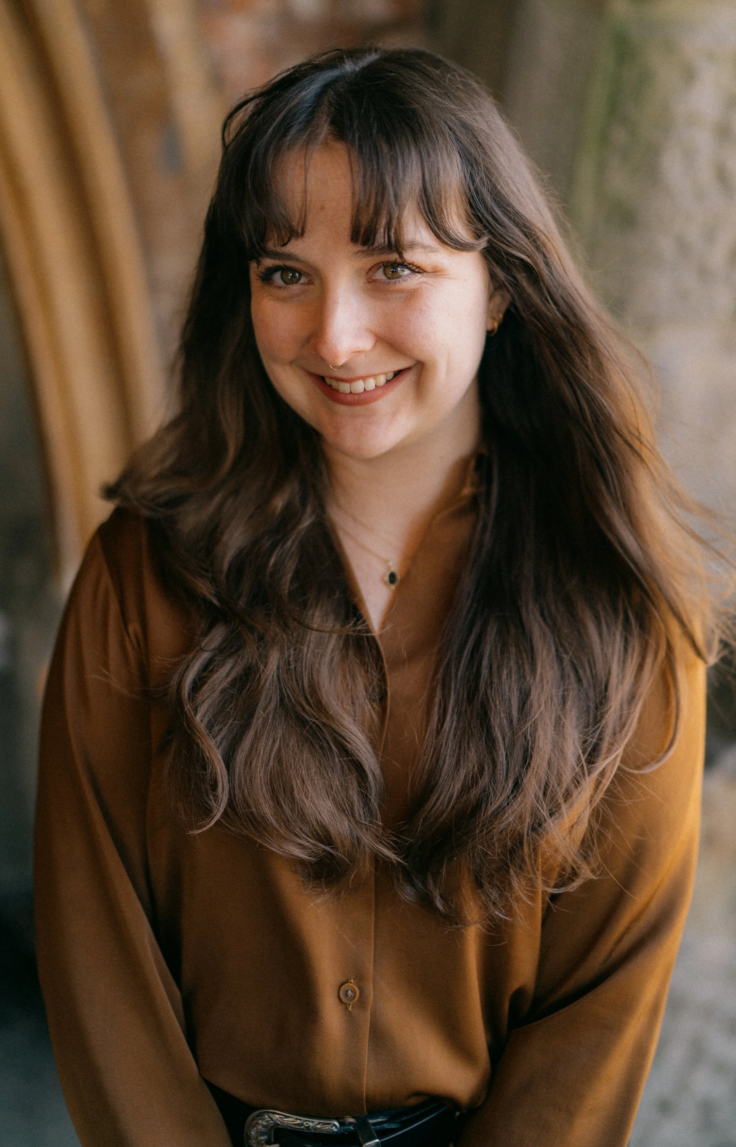 A woman with long wavy brown hair and brown eyes, smiling, wearing a brown shirt, standing indoors with a brick wall in the background.