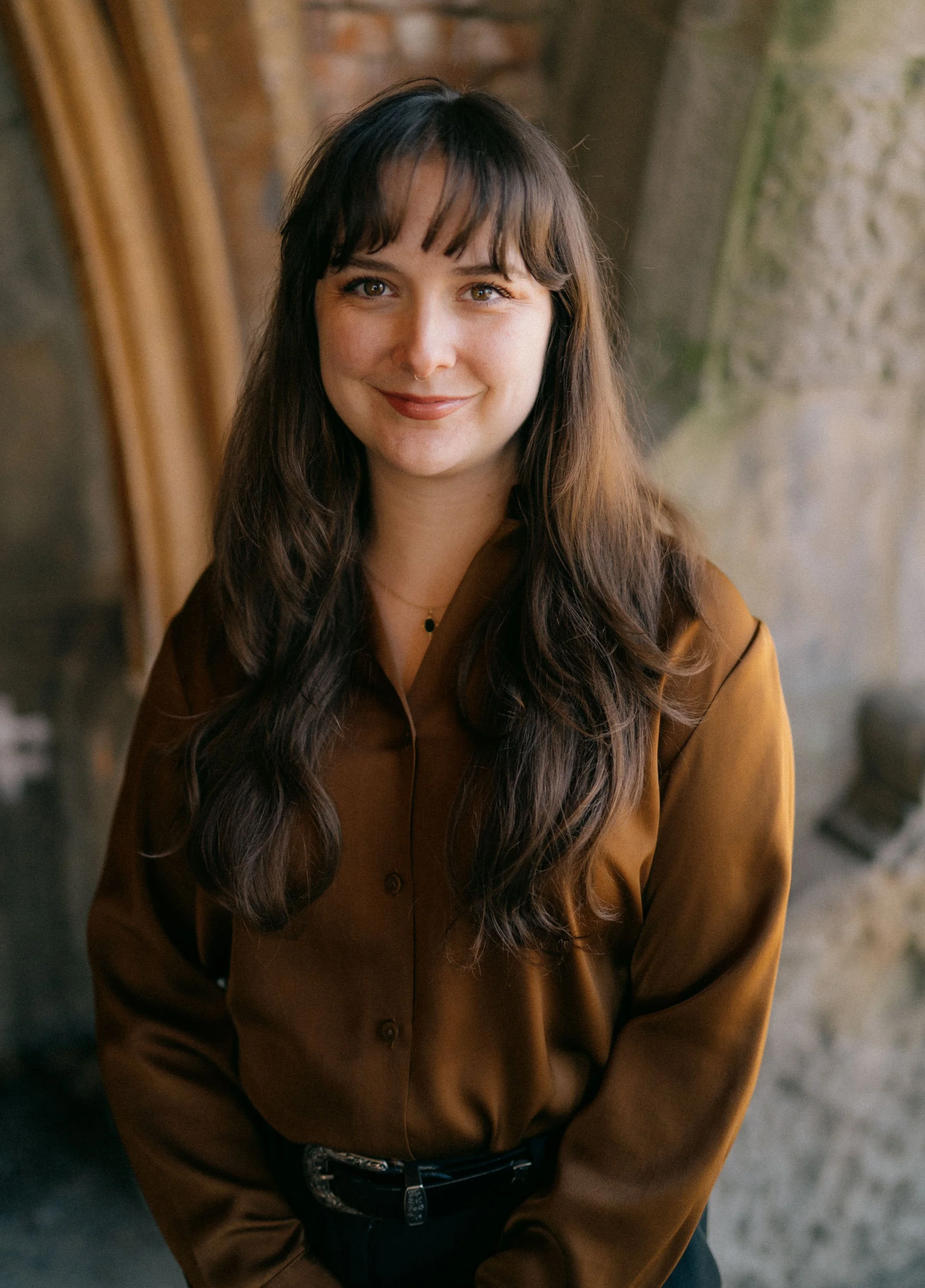 A woman with long wavy brown hair and bangs smiling, dressed in a brown button-up shirt, standing indoors near a brick and concrete wall.