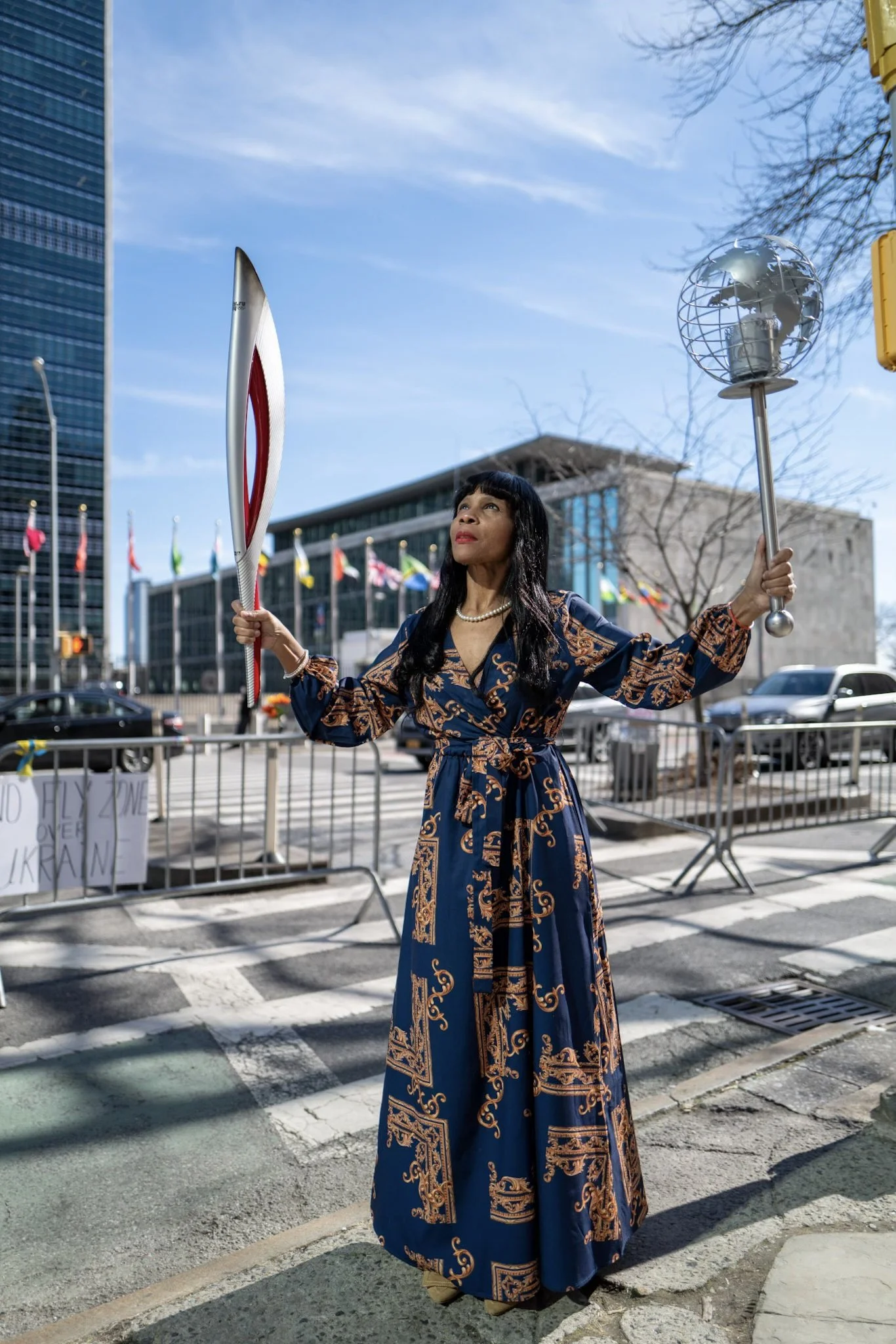 A woman standing on a city street holding a mirror and a lamp while looking upward. She is dressed in a long, patterned navy and gold dress and wears a pearl necklace. There are flags in the background and metal barriers on the sidewalk.