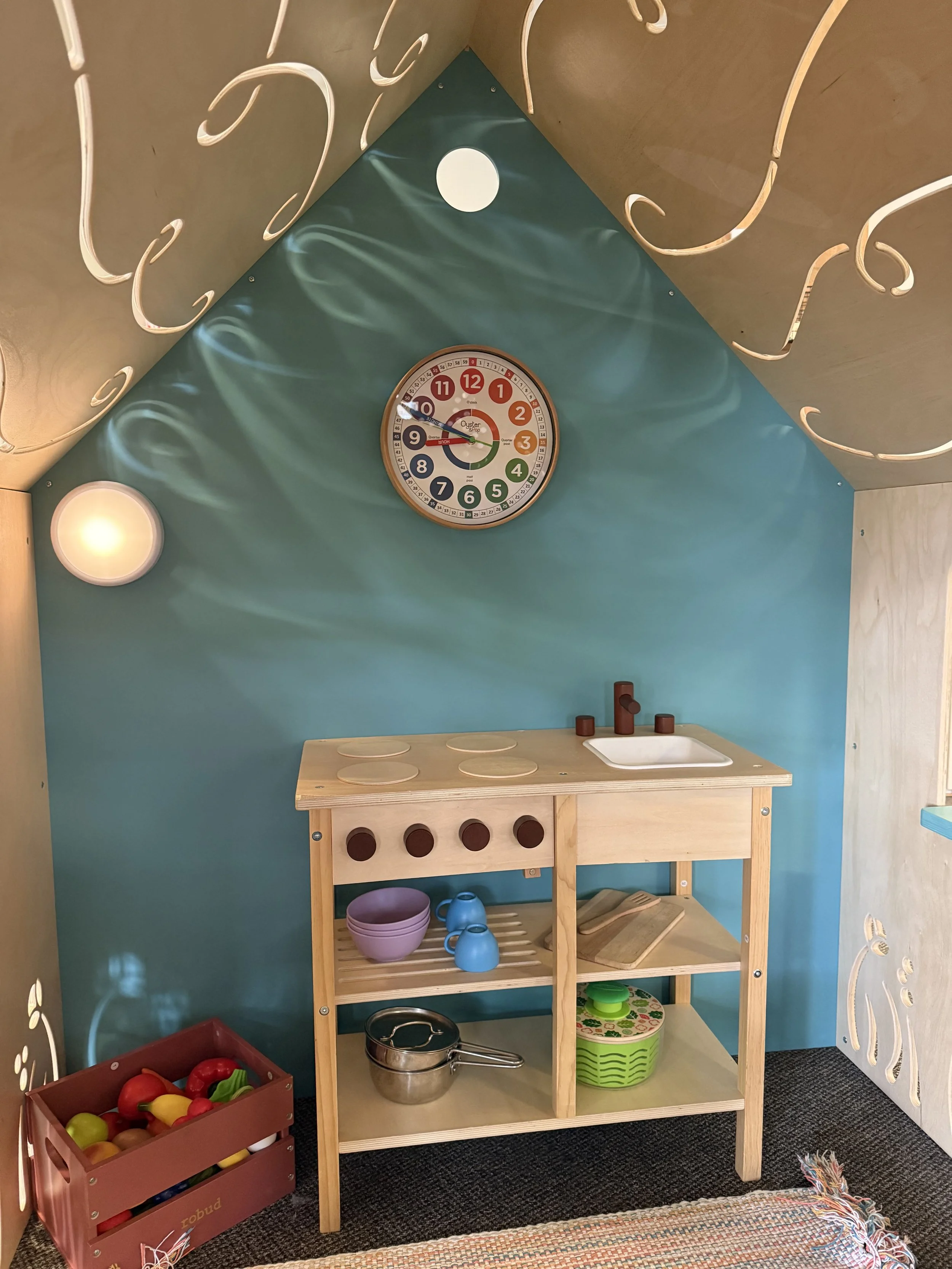 A children's pretend play kitchen with a blue wall, wooden table, and toy kitchen accessories, including a small sink and stovetop, in a cozy room with colorful decor.