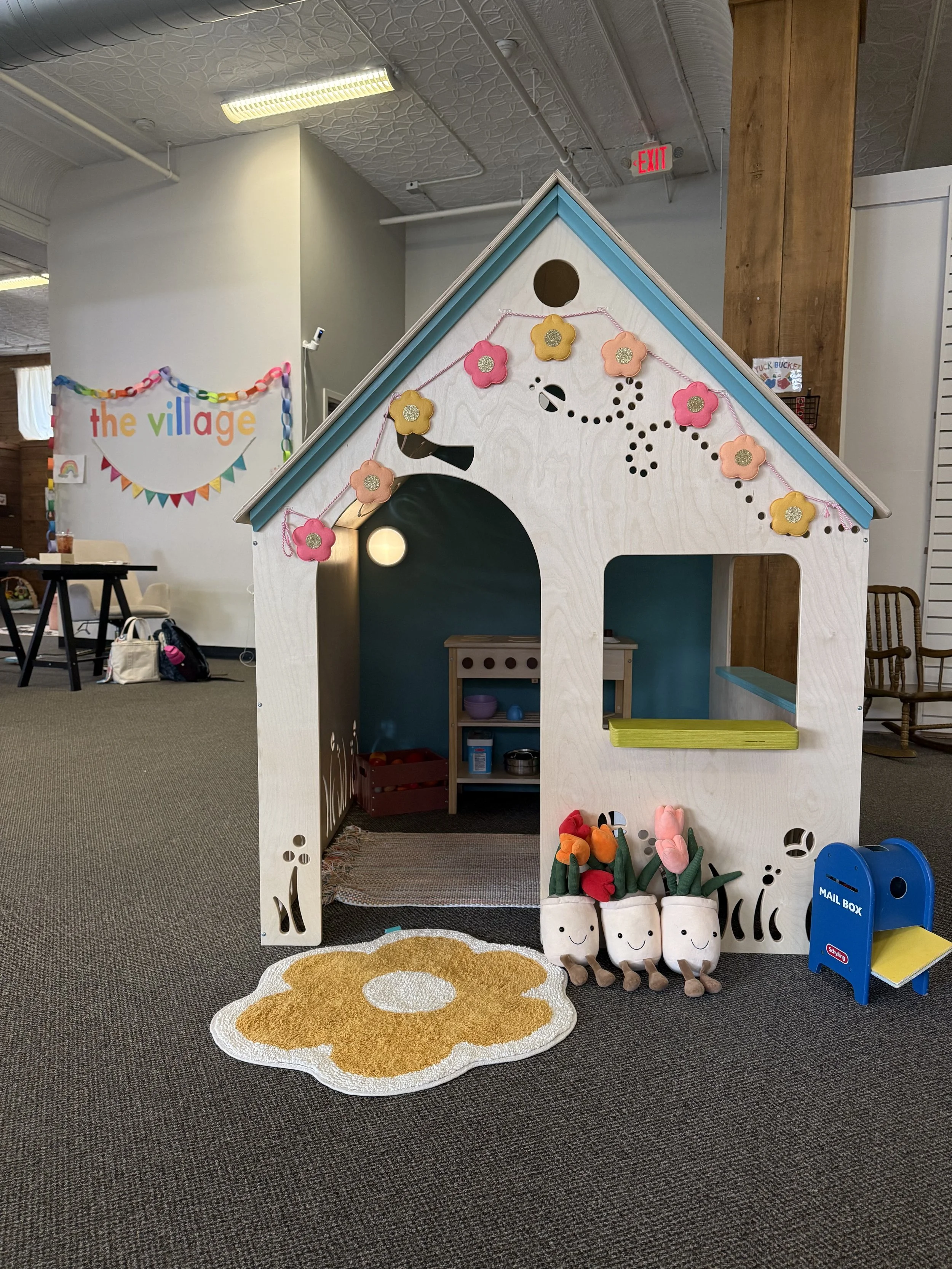 Children's playhouse decorated with felt flowers and a string of pink flowers, situated indoors with a rug shaped like a flower in front and plush plants with smiling faces nearby.