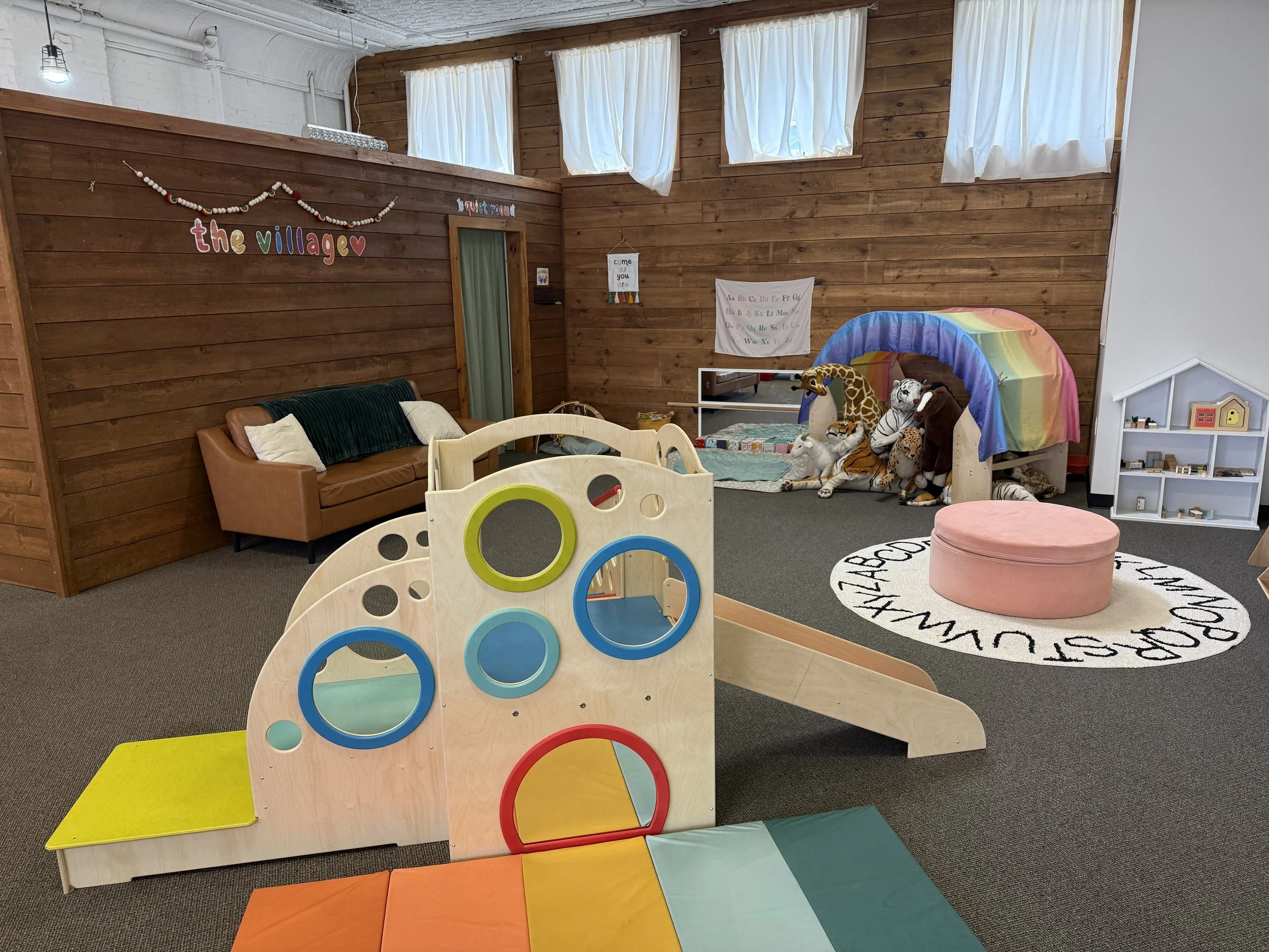 Interior of a children's playroom with wooden walls, a brown couch with pillows, a colorful play structure with circular openings, plush toys including a giraffe, zebra, and elephant under a rainbow, a pink round ottoman, and various educational decorations on the walls.