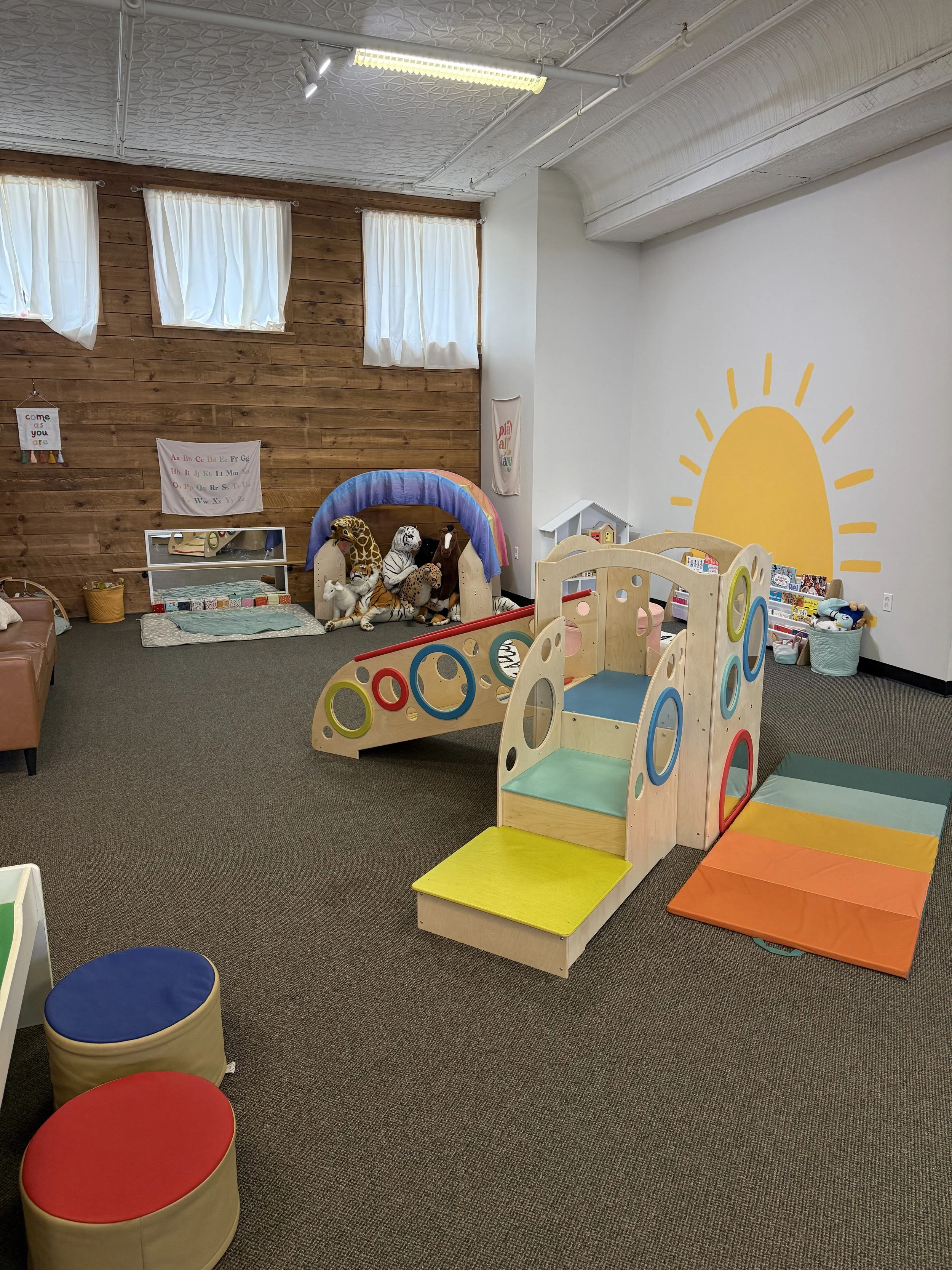 Children's indoor playroom with colorful playground equipment, stuffed animal toys, and a wall mural of a yellow sun on a white wall, with wooden wall panels and windows with white curtains.
