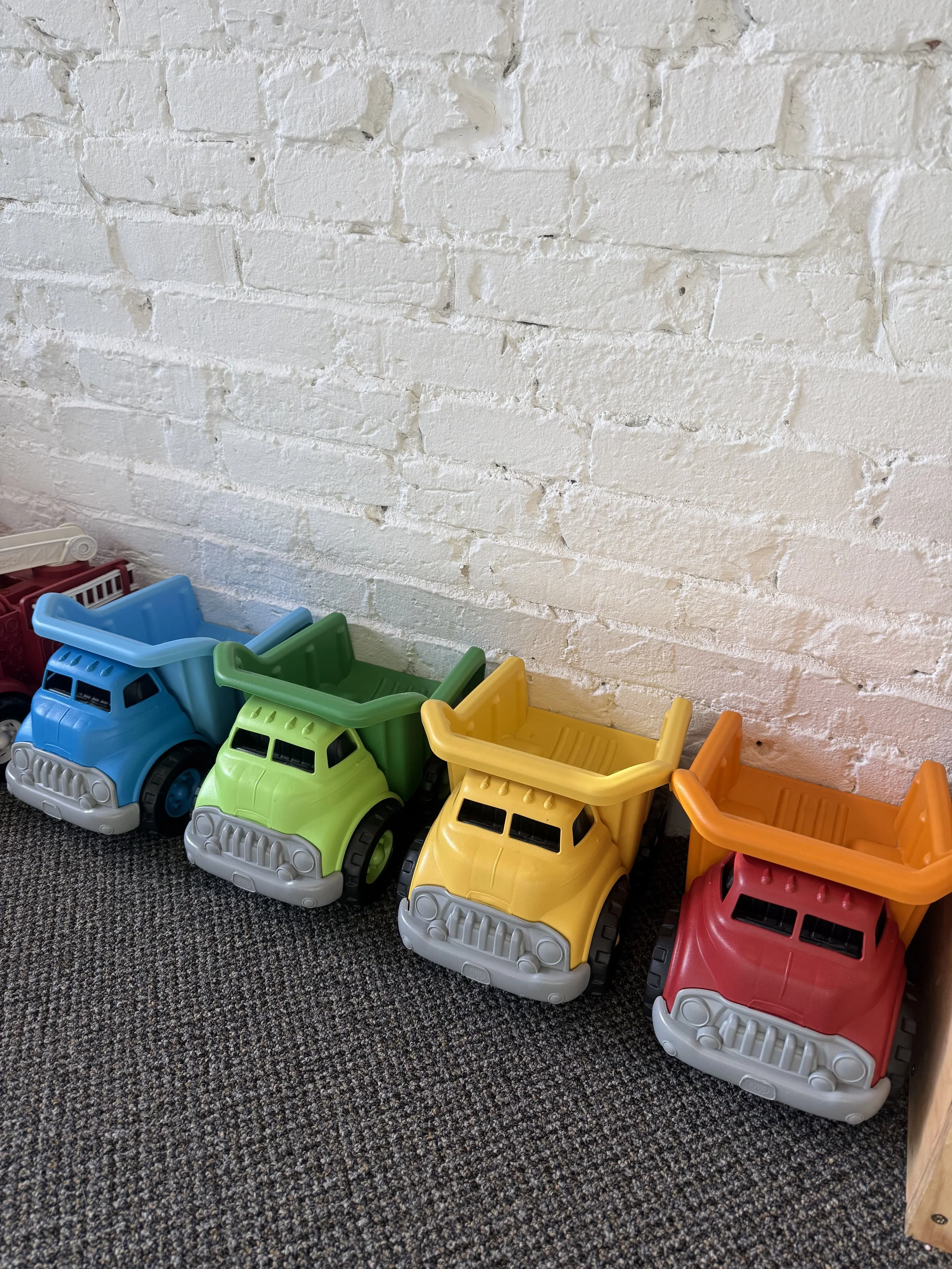 Colorful toy dump trucks lined up against a white brick wall on a gray carpet.