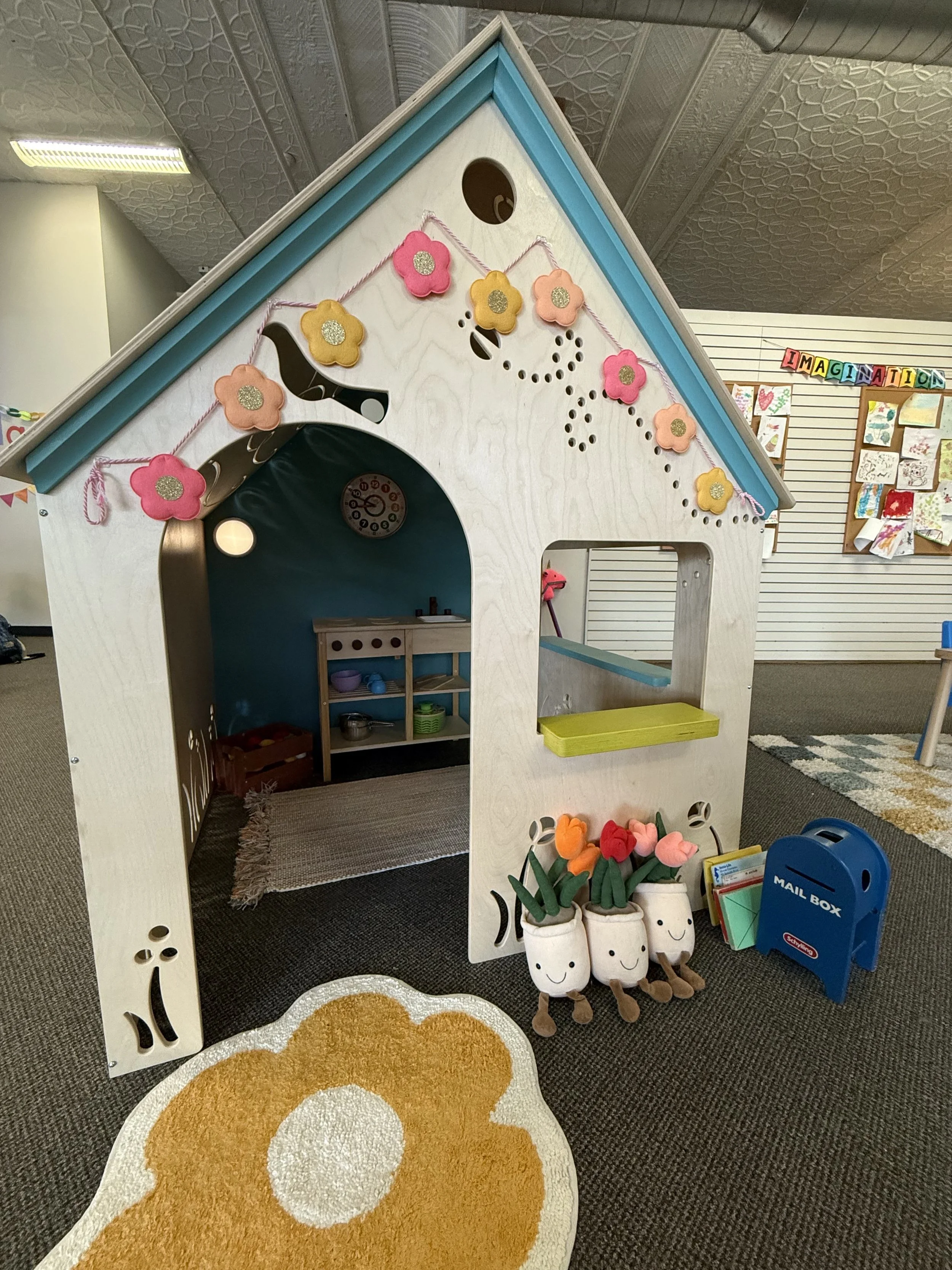 Children's playhouse with flower decorations, a mailbox, and plush pots labeled 'M' and 'N' on a carpeted floor.