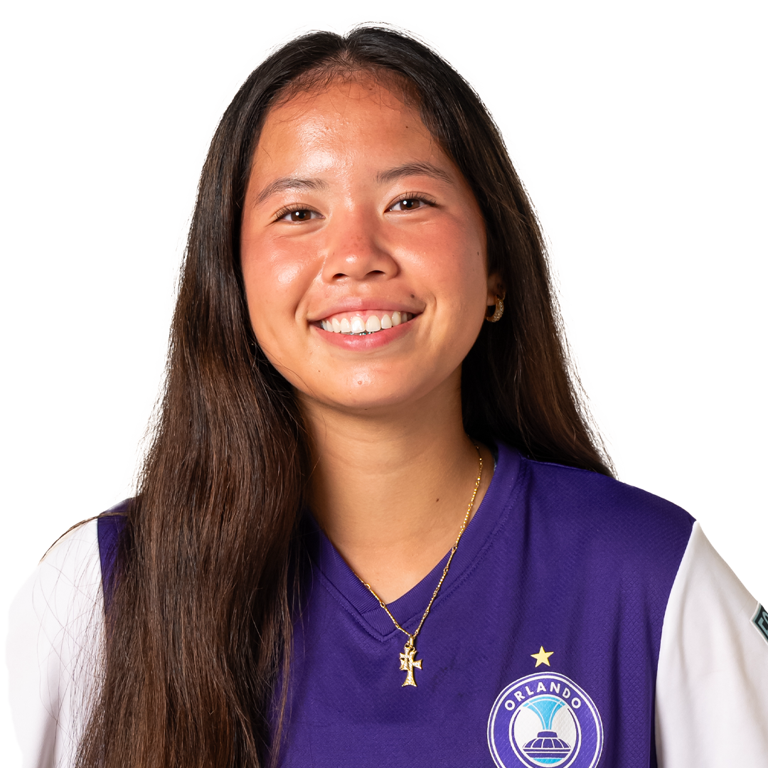 A young woman with long brown hair, smiling, wearing a purple Orlando soccer jersey with a white emblem and a gold cross necklace.