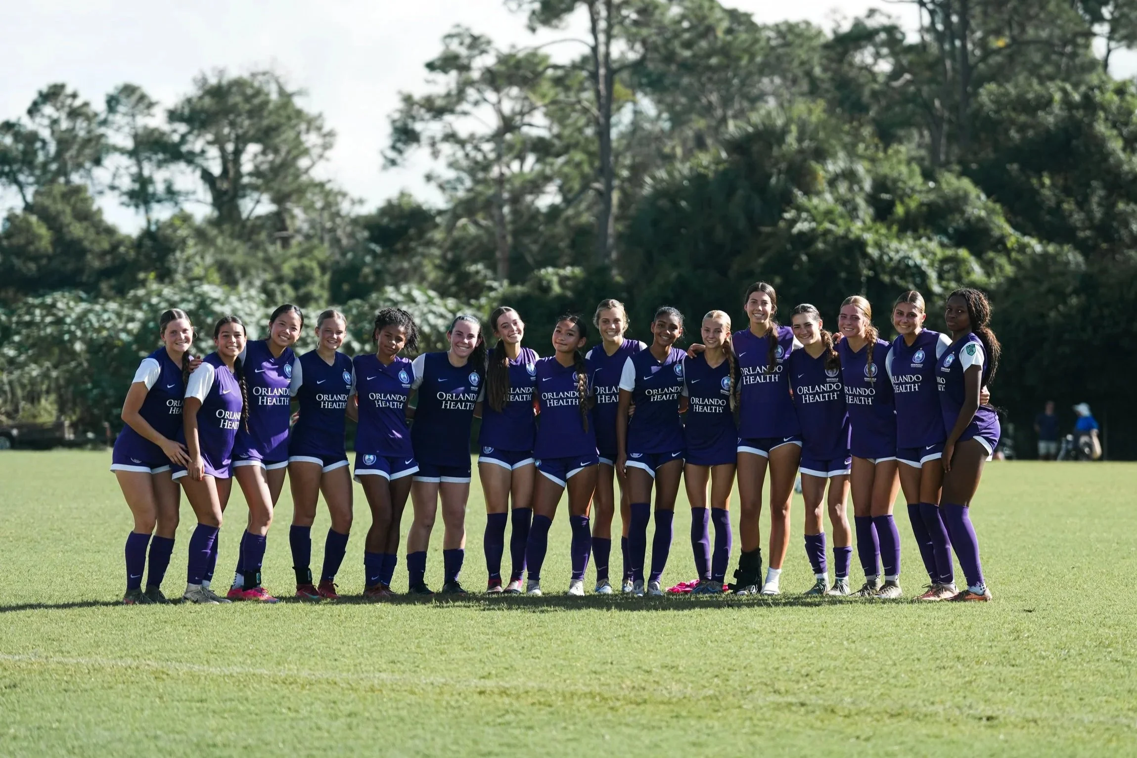 Group of women soccer players standing on a field, smiling, wearing purple and white uniforms with 'Orlando Health' logo.