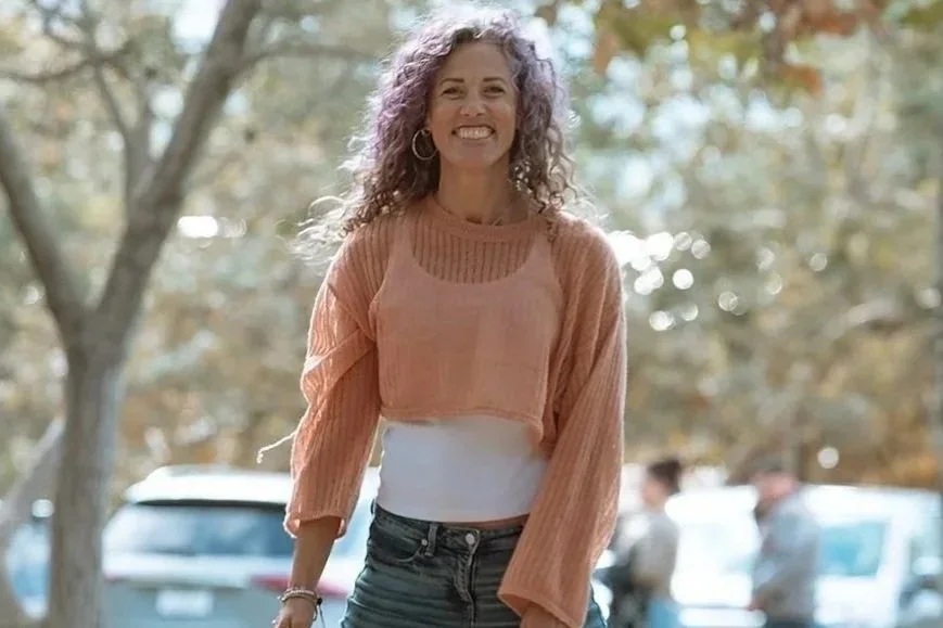 A woman outdoors smiling with trees and cars in the background.