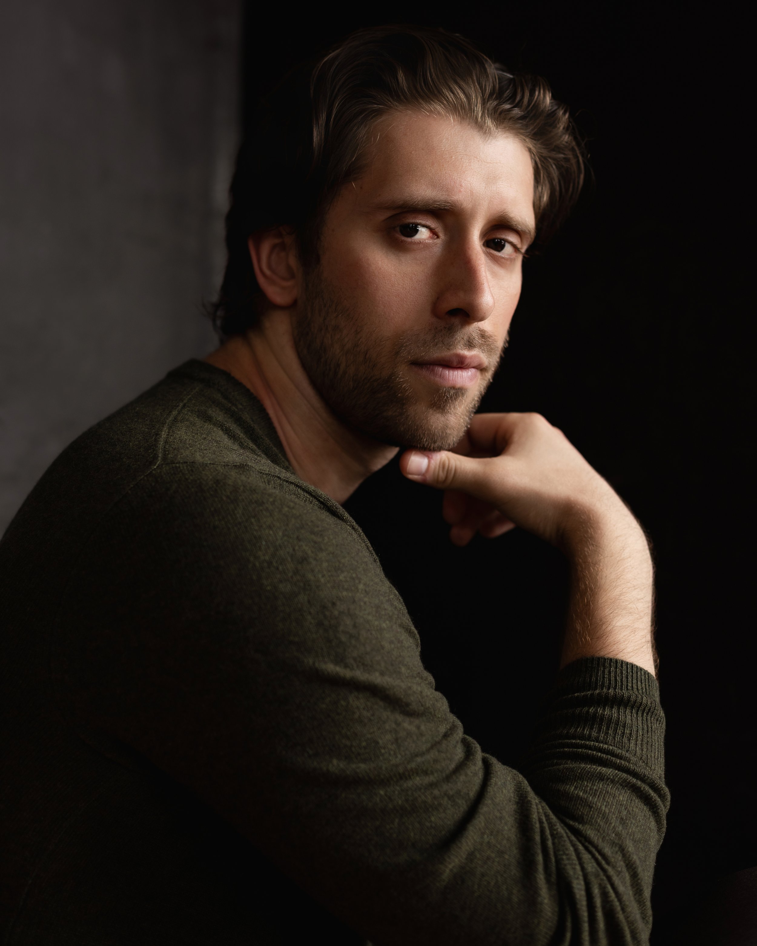 A portrait of a young man with brown hair and a beard, wearing a dark green sweater, against a black background.