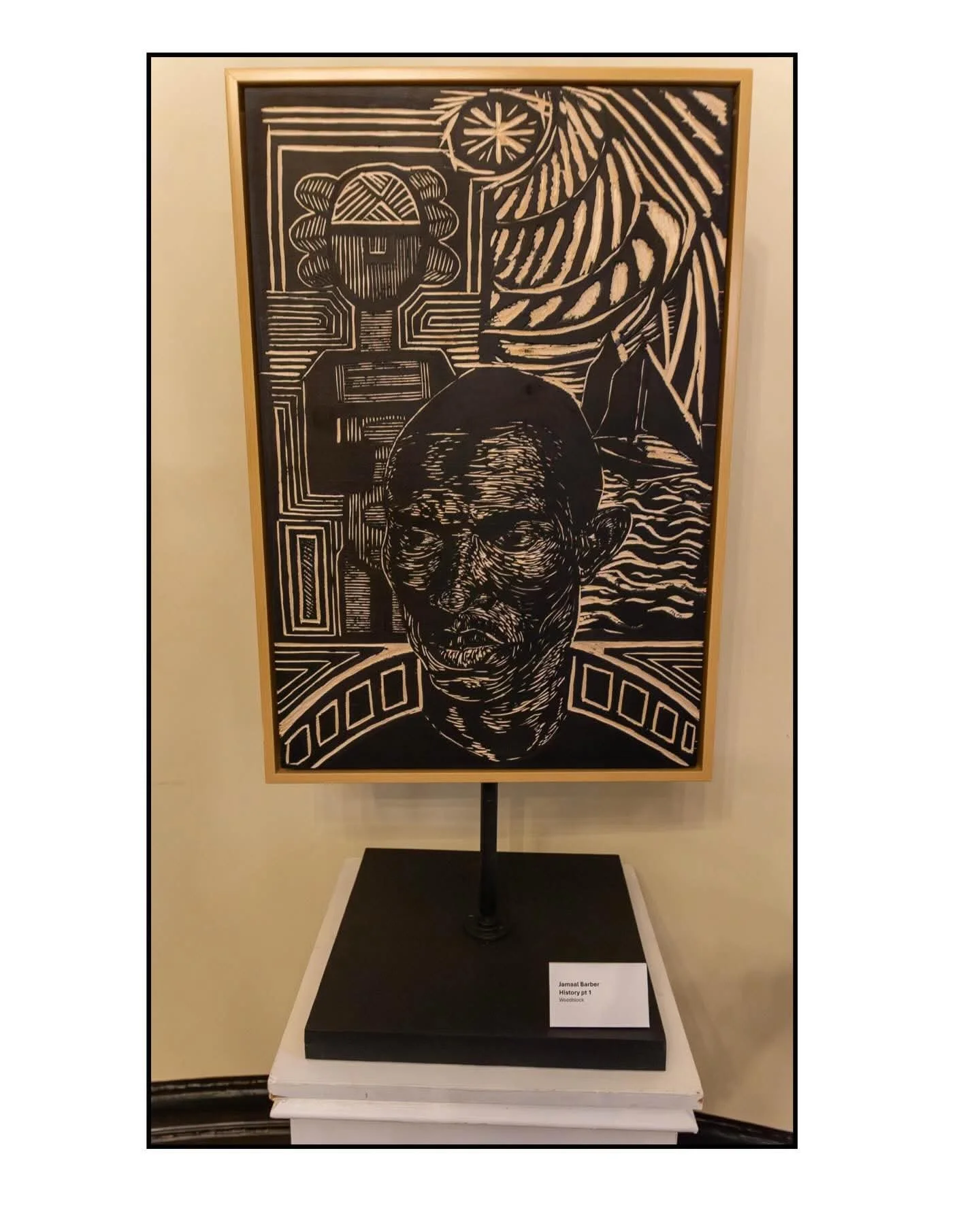 A man sewing on a sewing machine at a table in a brightly lit room, with an elderly woman smiling behind him. The table is decorated with a quilt. There are colorful craft projects and a window with curtains in the background.