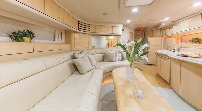 Interior of a luxurious yacht with a curved cream-colored sofa, beige cushions, a wooden table with a vase of green leaves, and a kitchenette with a sink and plants in the background.