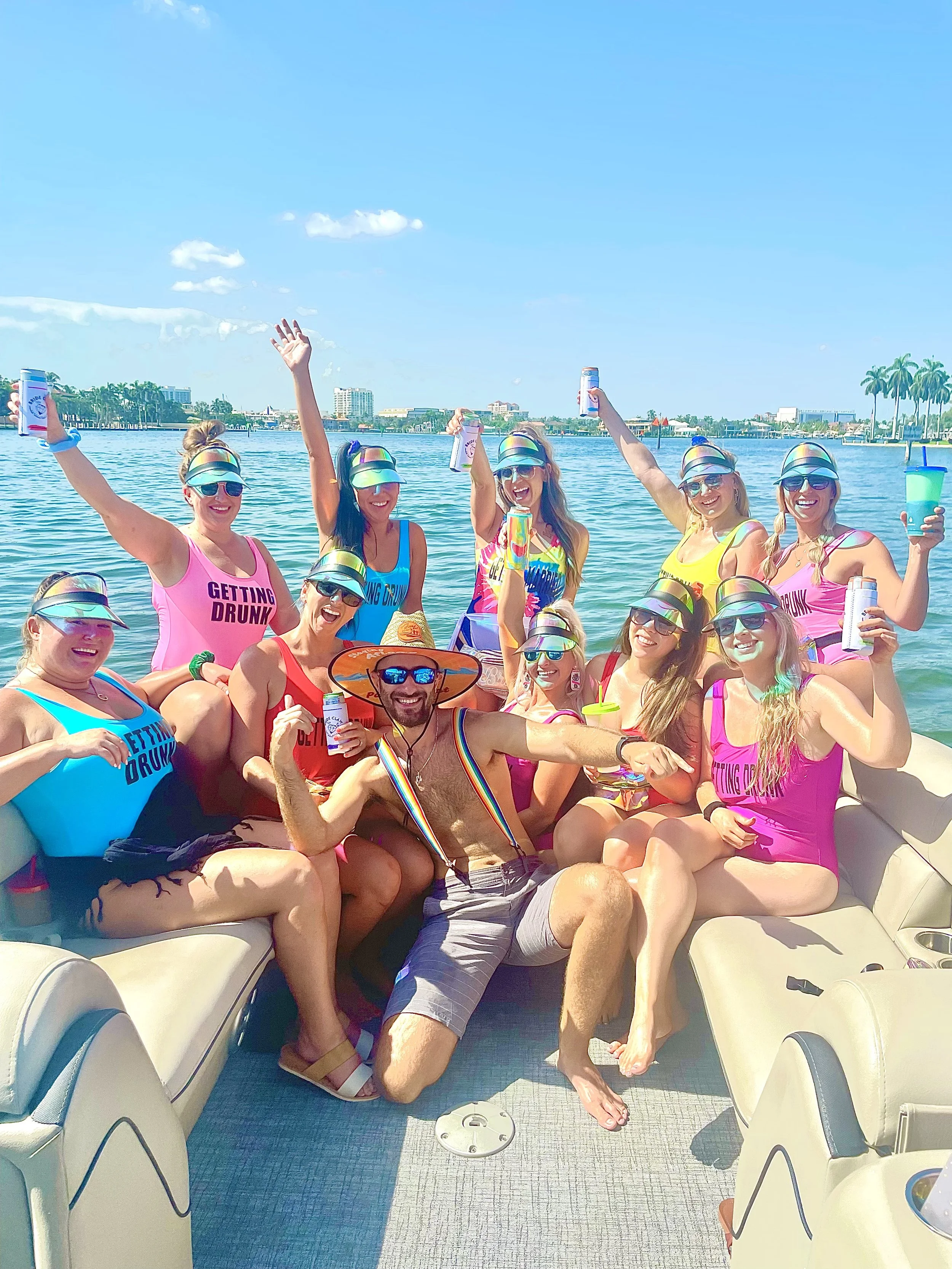 Group of people celebrating on a boat in a marina, with water and city skyline in the background, wearing colorful swimsuits, hats, and sunglasses, holding drinks, with one person wearing a hat and suspenders.