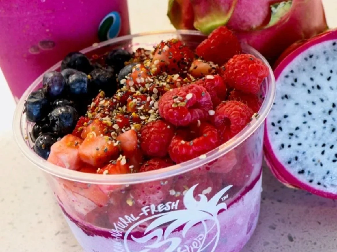 A clear plastic cup filled with layered yogurt and topped with sliced strawberries, blueberries, raspberries, and drizzled with chia seeds. Surrounding the cup are dragon fruit and other fruits.