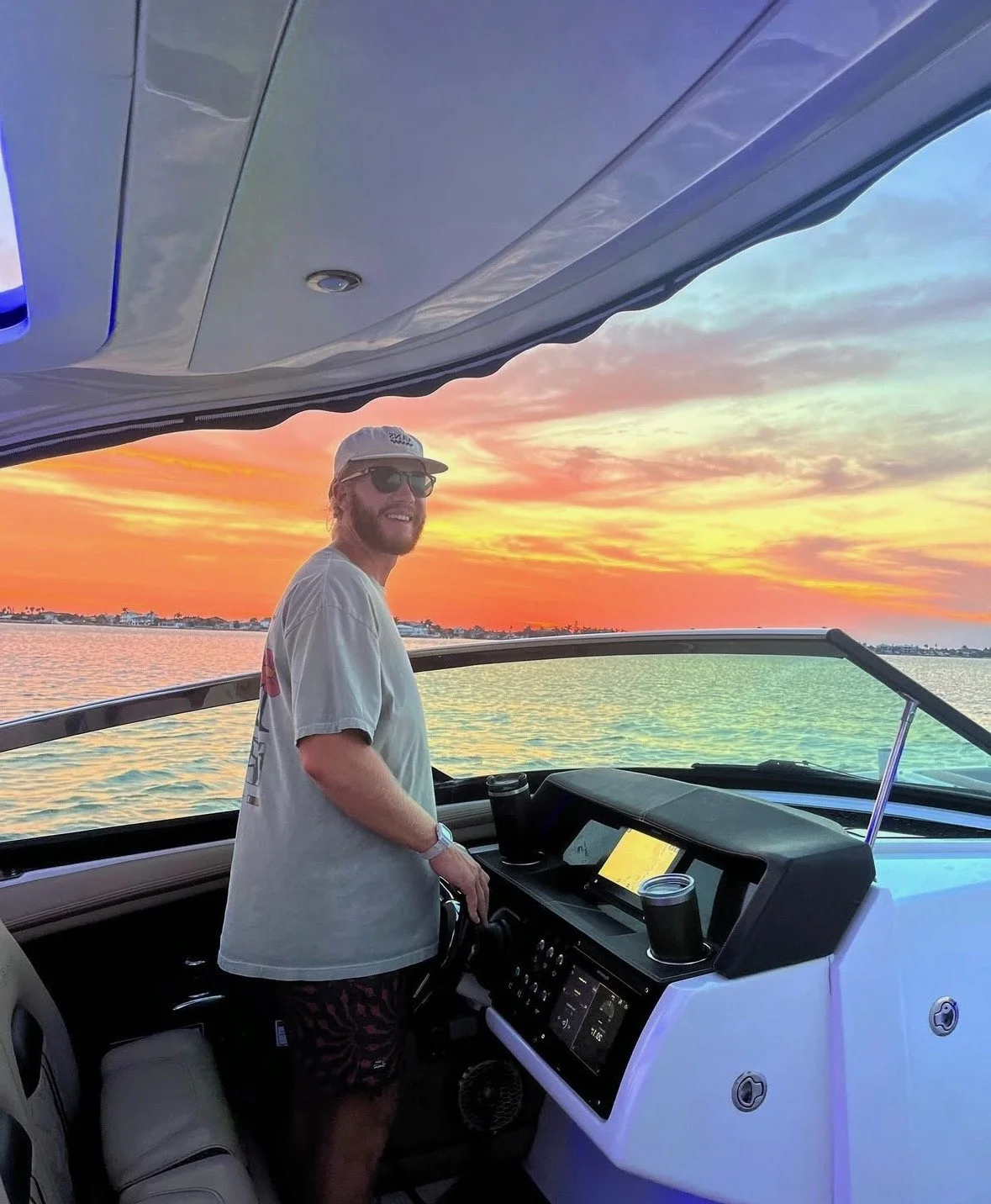 A man wearing sunglasses, a cap, and a t-shirt is steering a boat during sunset on the water, with colorful sky and clouds in the background in Fort Lauderdale .