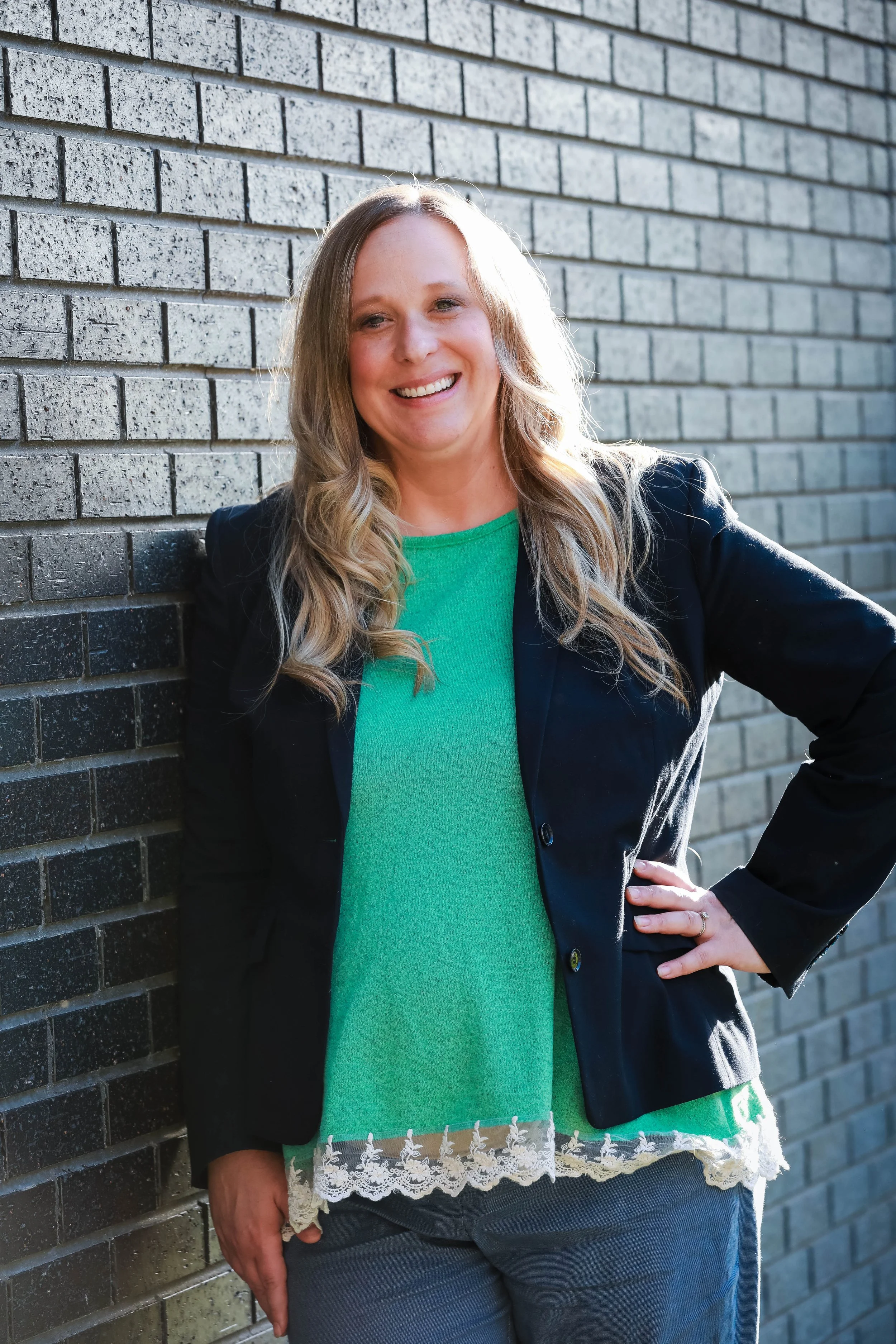 A woman with long wavy blonde hair, smiling and standing with one hand on her hip, leaning against a brick wall outdoors.