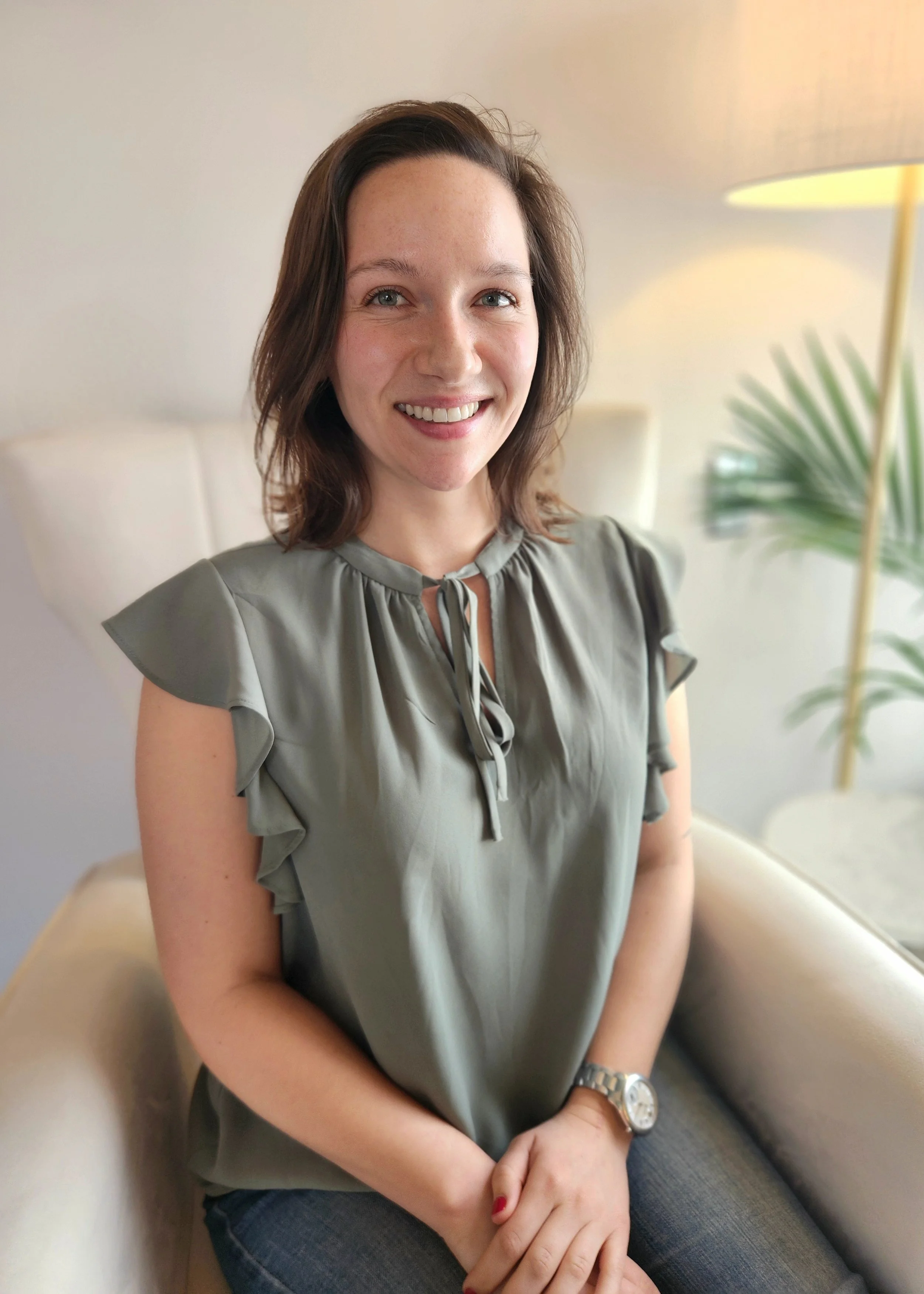 A woman with shoulder-length brown hair, wearing a gray blouse with ruffled sleeves and a watch, sitting on a beige chair in a well-lit room with a plant and lamp in the background.
