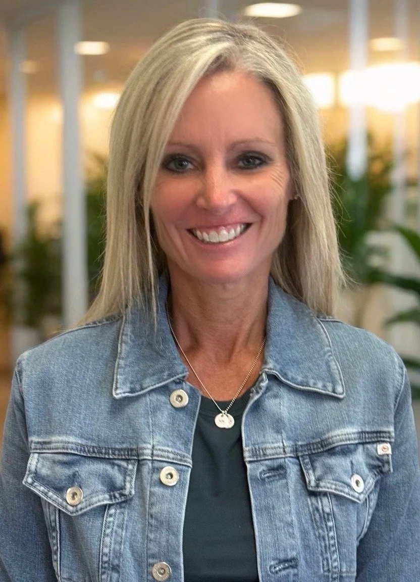 A woman with long blonde hair, wearing a beige jacket over a white top, smiling in an indoor setting with plants and warm lighting in the background.