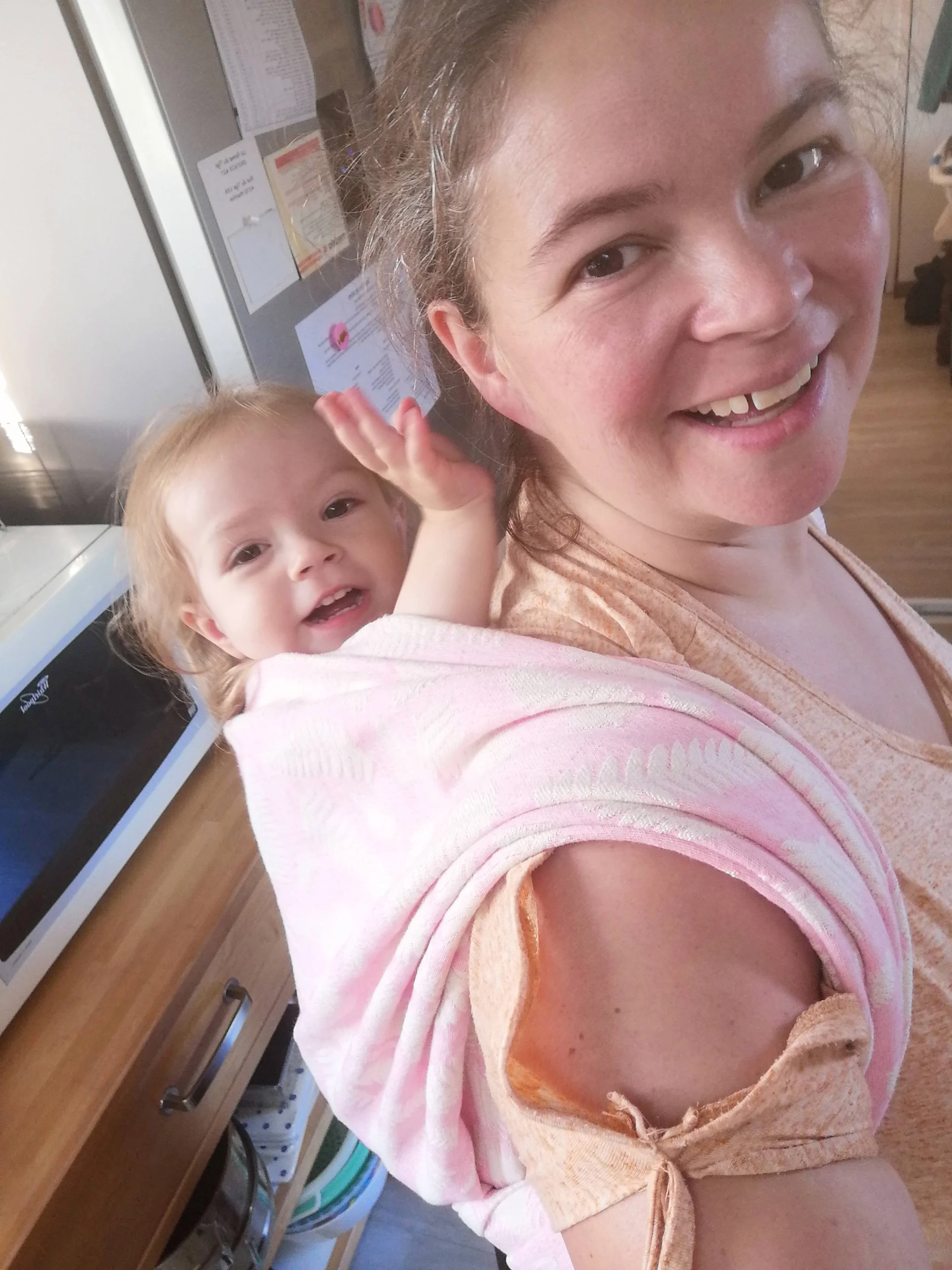Une femme souriante porte un bébé sur ses épaules, dans une cuisine, près d’un réfrigérateur.