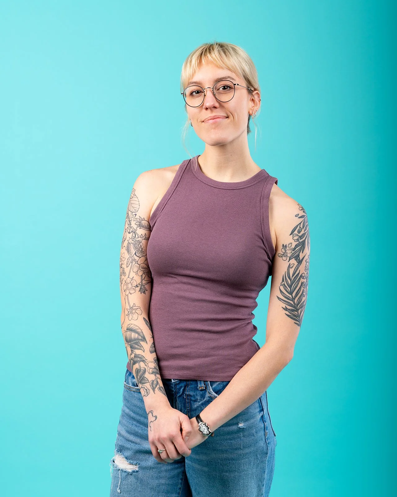 A woman with short blonde hair, glasses, and tattoos on her arms, wearing a sleeveless purple top and ripped jeans, standing against a light blue background.