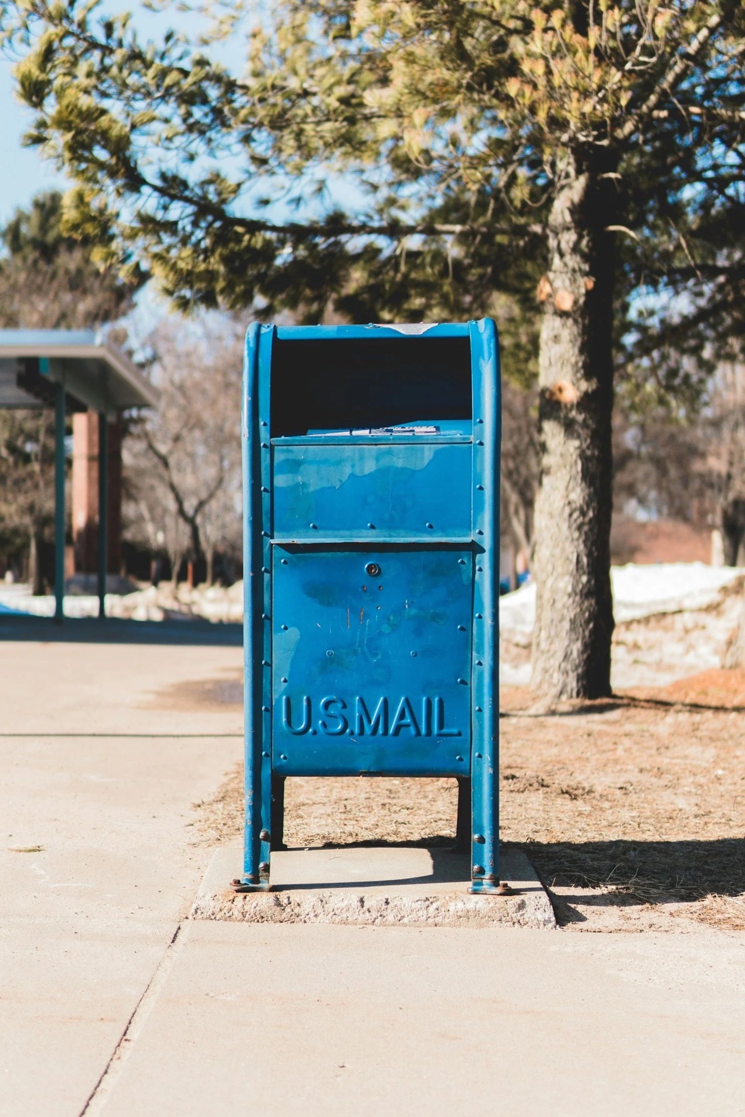A blue U.S. Mail collection box on a sidewalk with trees and a building in the background.
