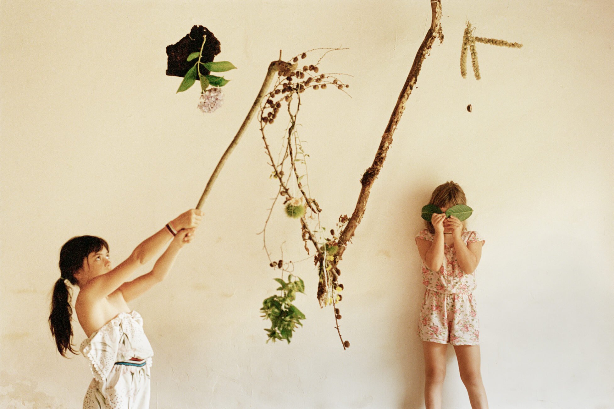 Zwei junge Mädchen spielen vor neutralem Hintergrund mit Zweigen und Blättern.