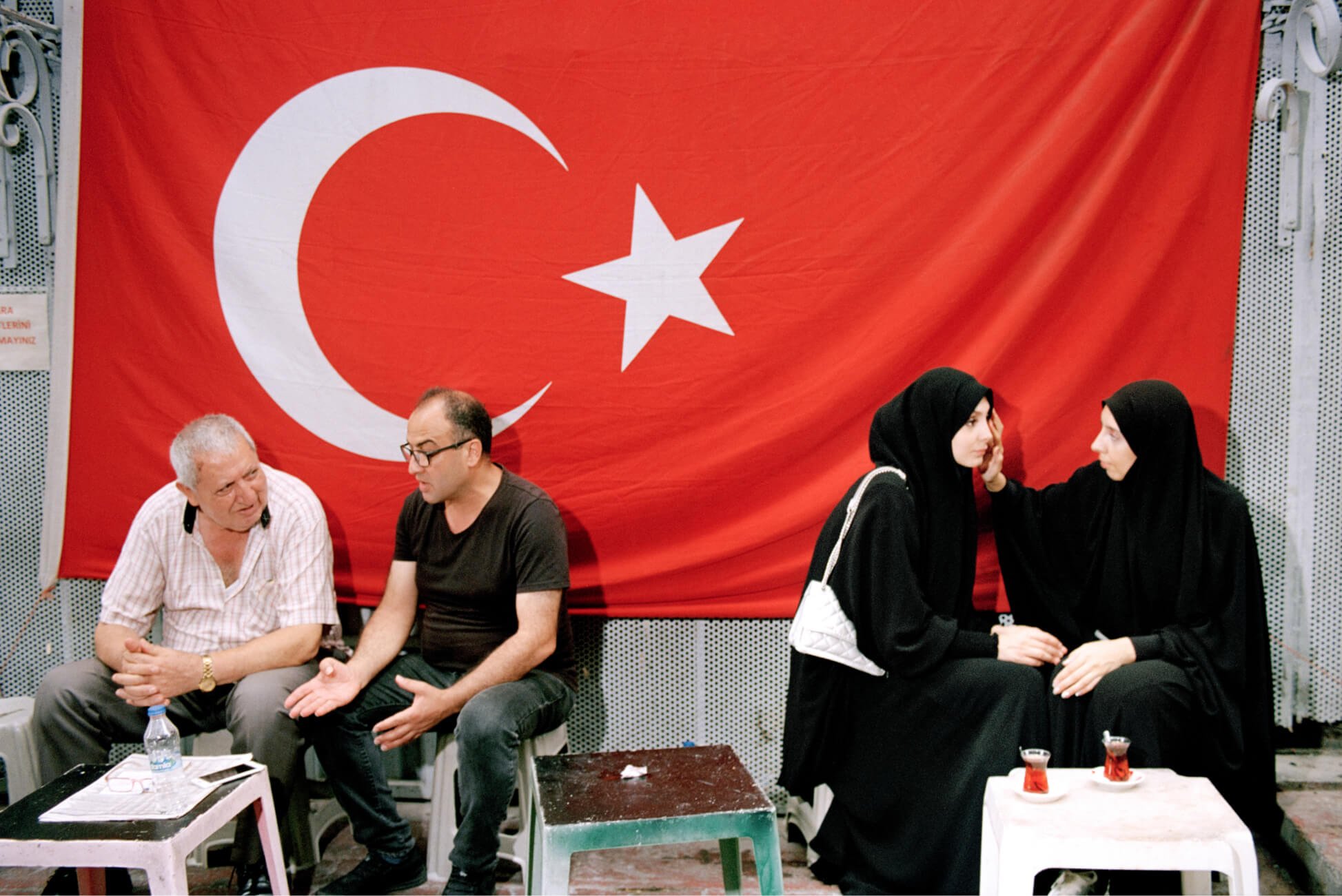 Zwei Männer und zwei verschleierte Frauen sitzen getrennt in einem Teeladen mit einer türkischen Flagge im Hintergrund.