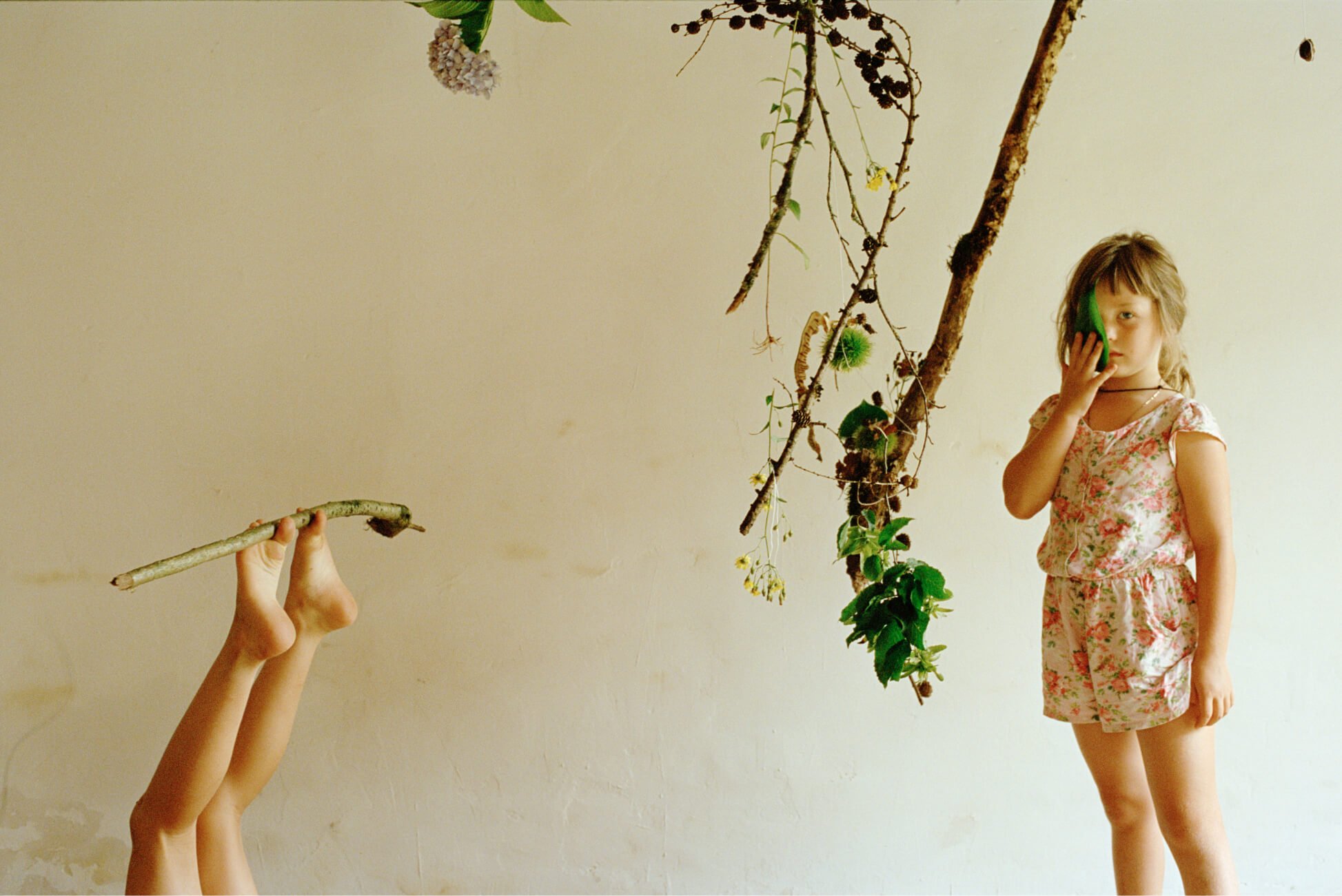 Une jeune fille tient une branche avec ses orteils tandis qu’une autre se couvre l’œil avec une grande feuille.