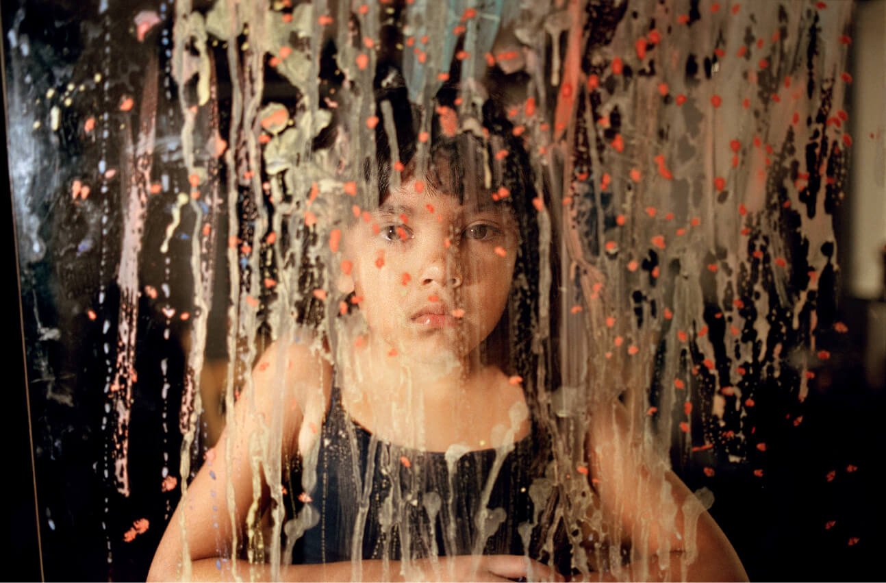 Une jeune fille regarde l’objectif à travers une vitre éclaboussée de peinture.
