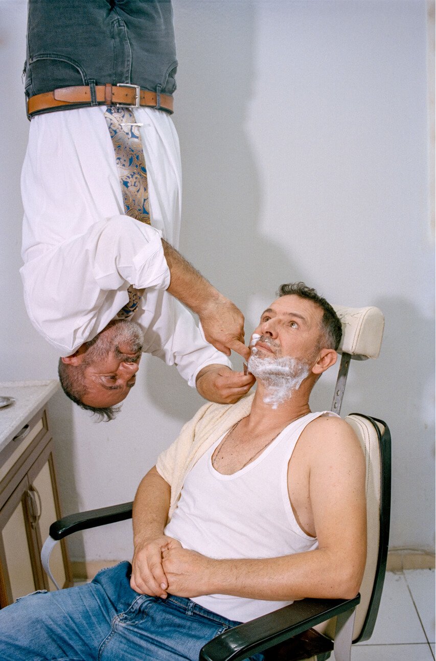Un hombre suspendido boca abajo afeita a otro hombre sentado en un sillón de barbería.