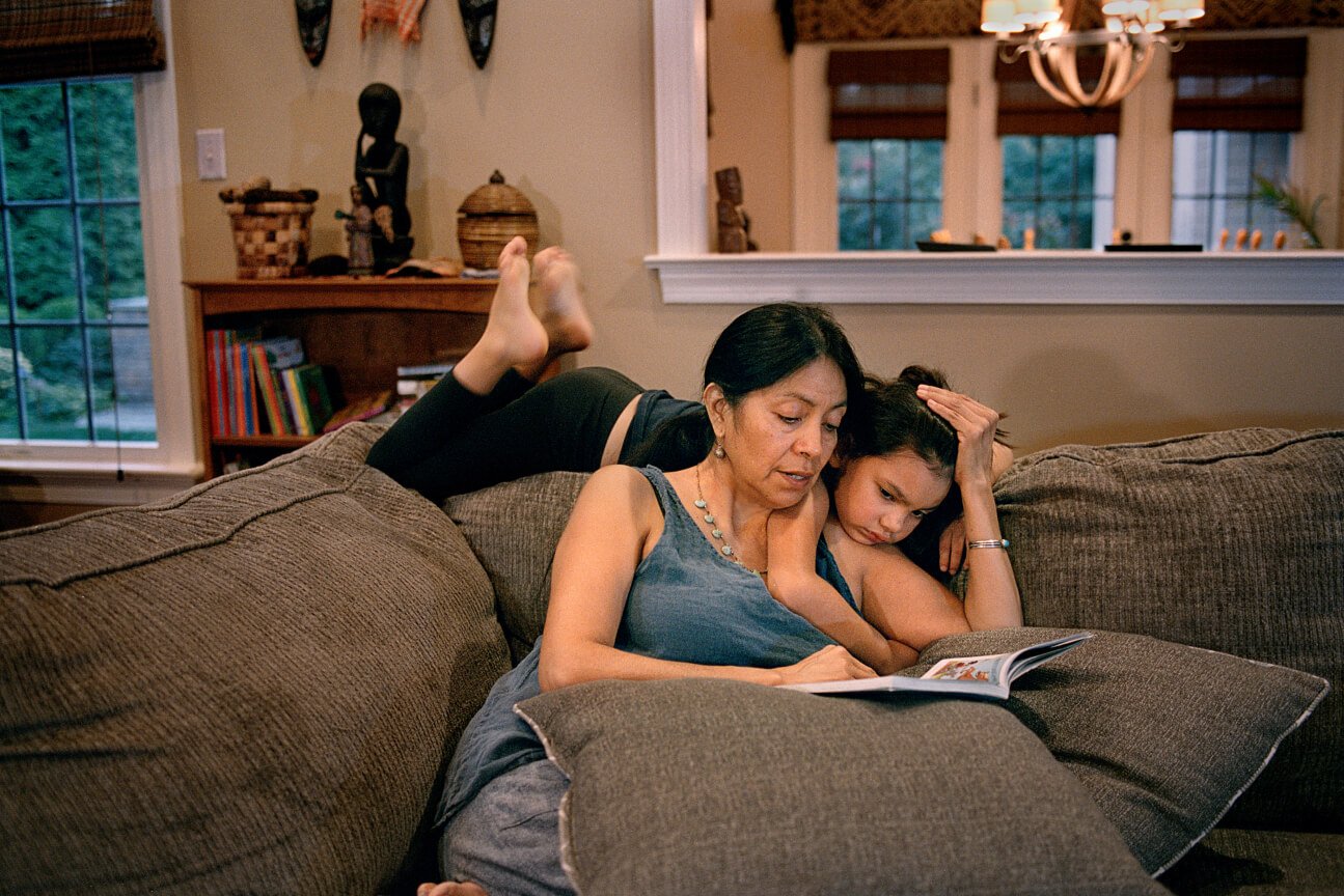 Mãe lê um livro para a filha deitada no encosto do sofá.