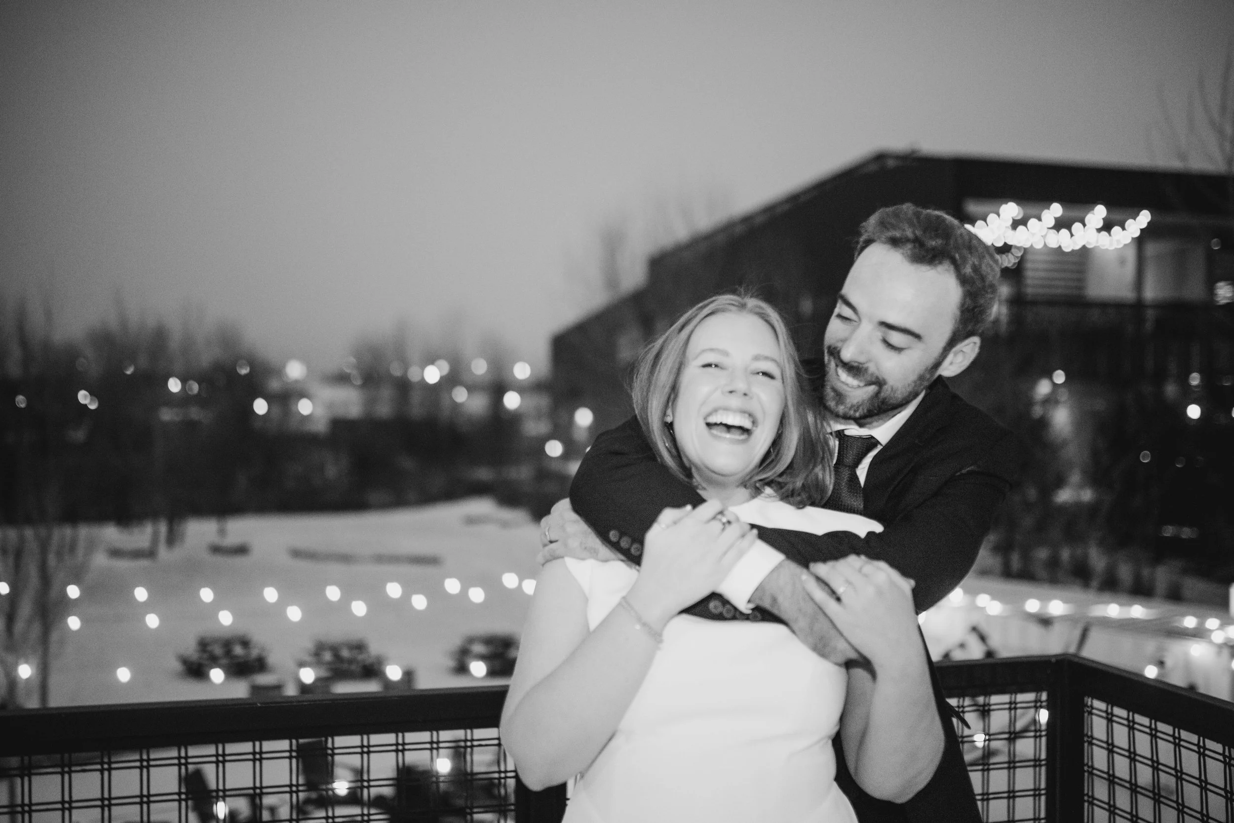 A couple dressed in formal attire sharing a joyful hug on a balcony at night with city lights and a building with string lights in the background