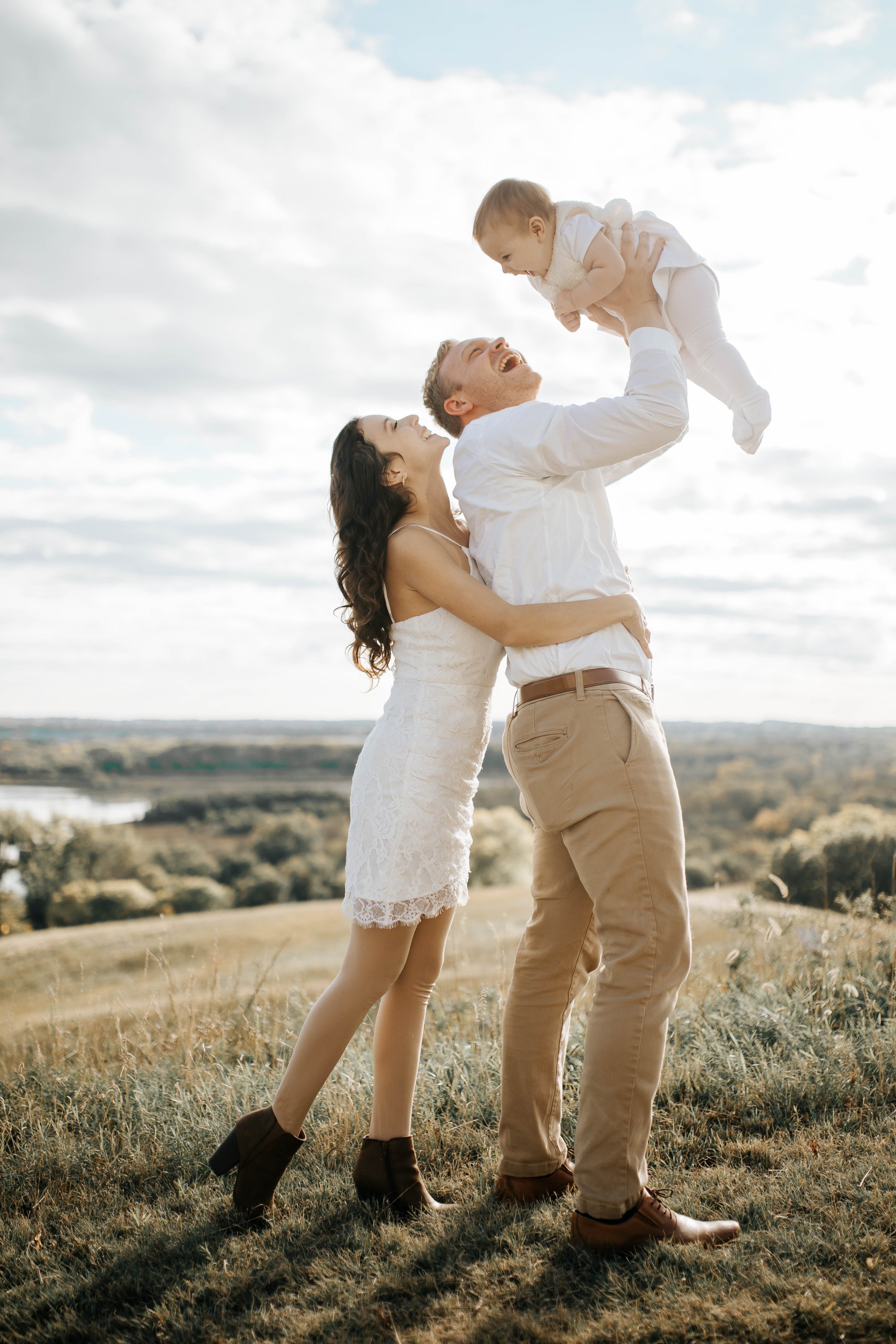A happy family outdoors on a grassy hill with a scenic background. The father, dressed in a white shirt and khaki pants, is lifting a baby girl dressed in white above his head. The mother, in a white lace dress, is standing close and smiling at them,