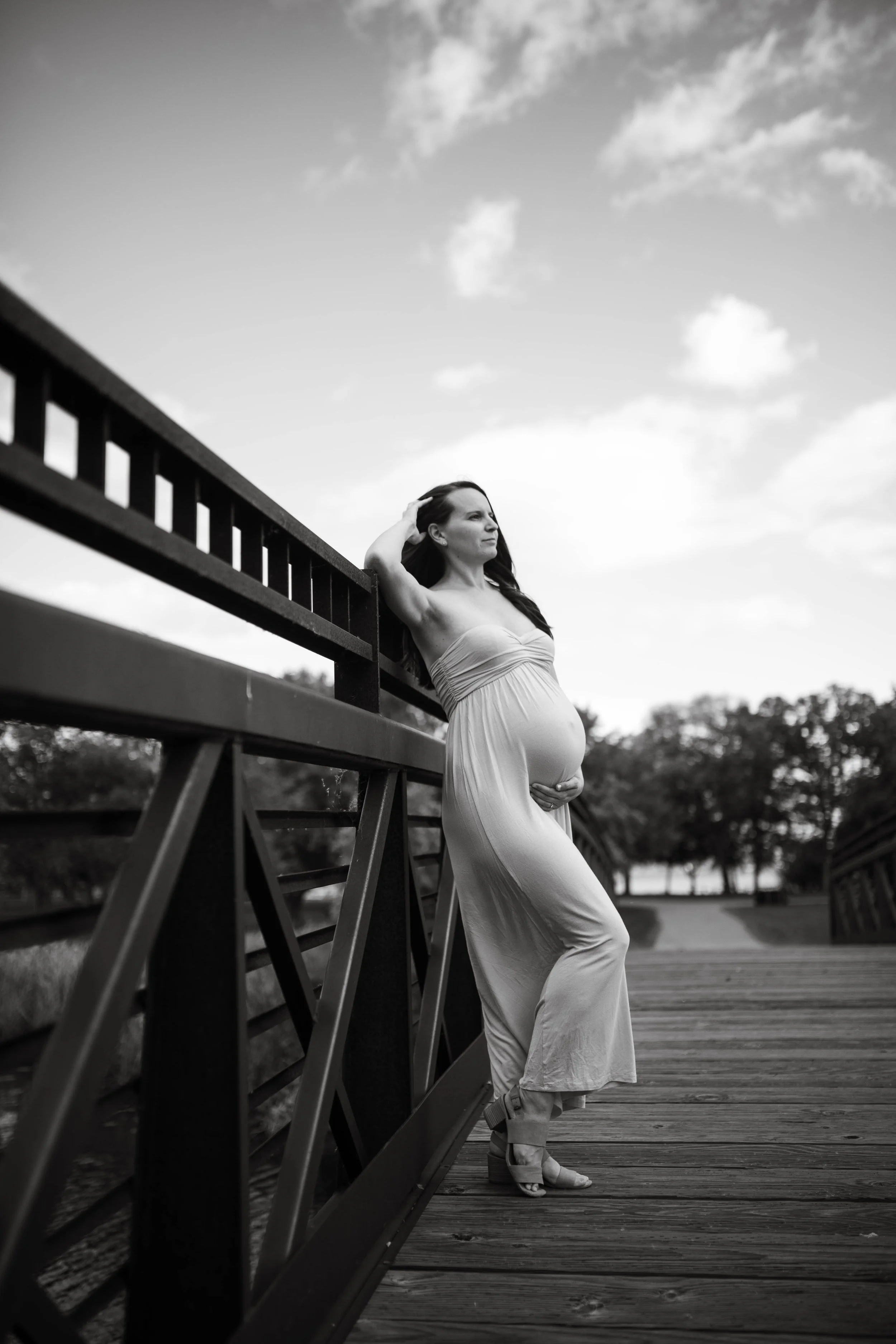 A pregnant woman in a strapless dress stands on a bridge, resting her back against the railing with a thoughtful expression. She is holding her belly and looking into the distance, with trees and a partly cloudy sky in the background.