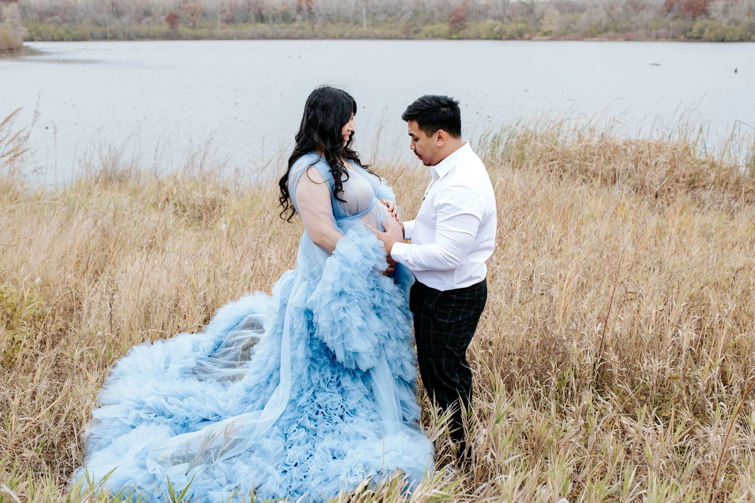 A couple standing in a field near a lake during autumn, with the man holding the pregnant woman's belly as she wears a flowing blue dress.