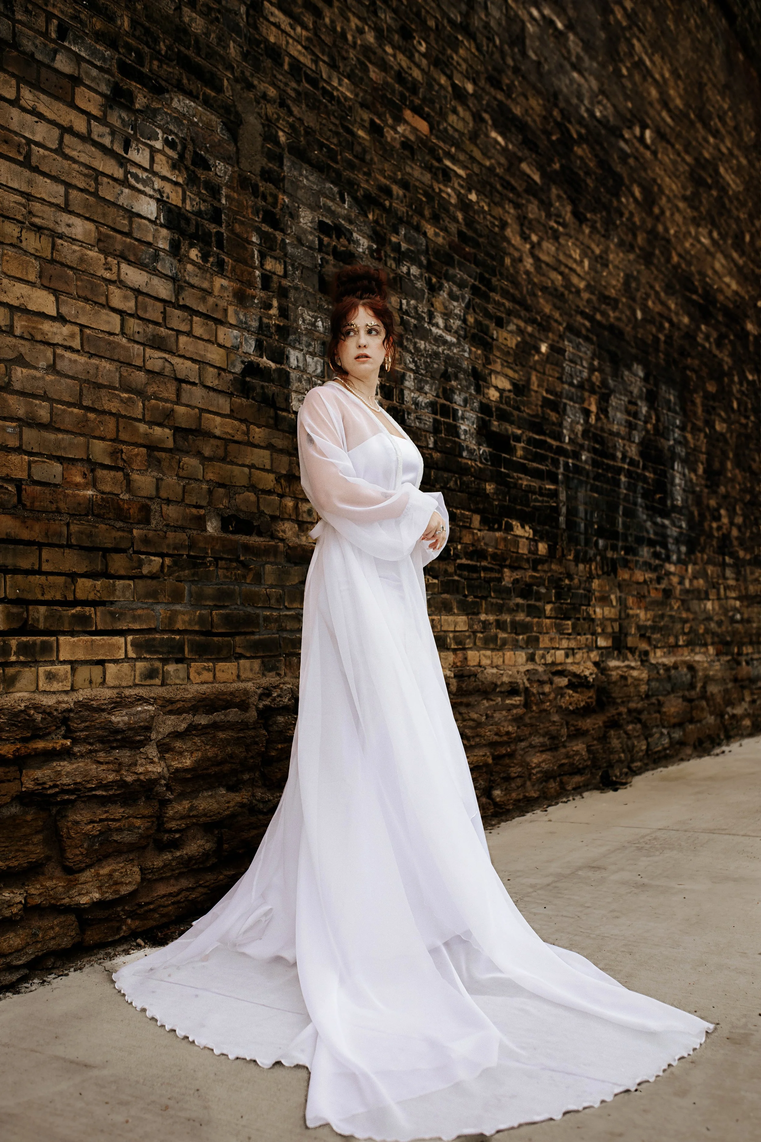 A woman wearing a long white dress with sheer sleeves stands against an old brick wall, looking to the side with a pensive expression.