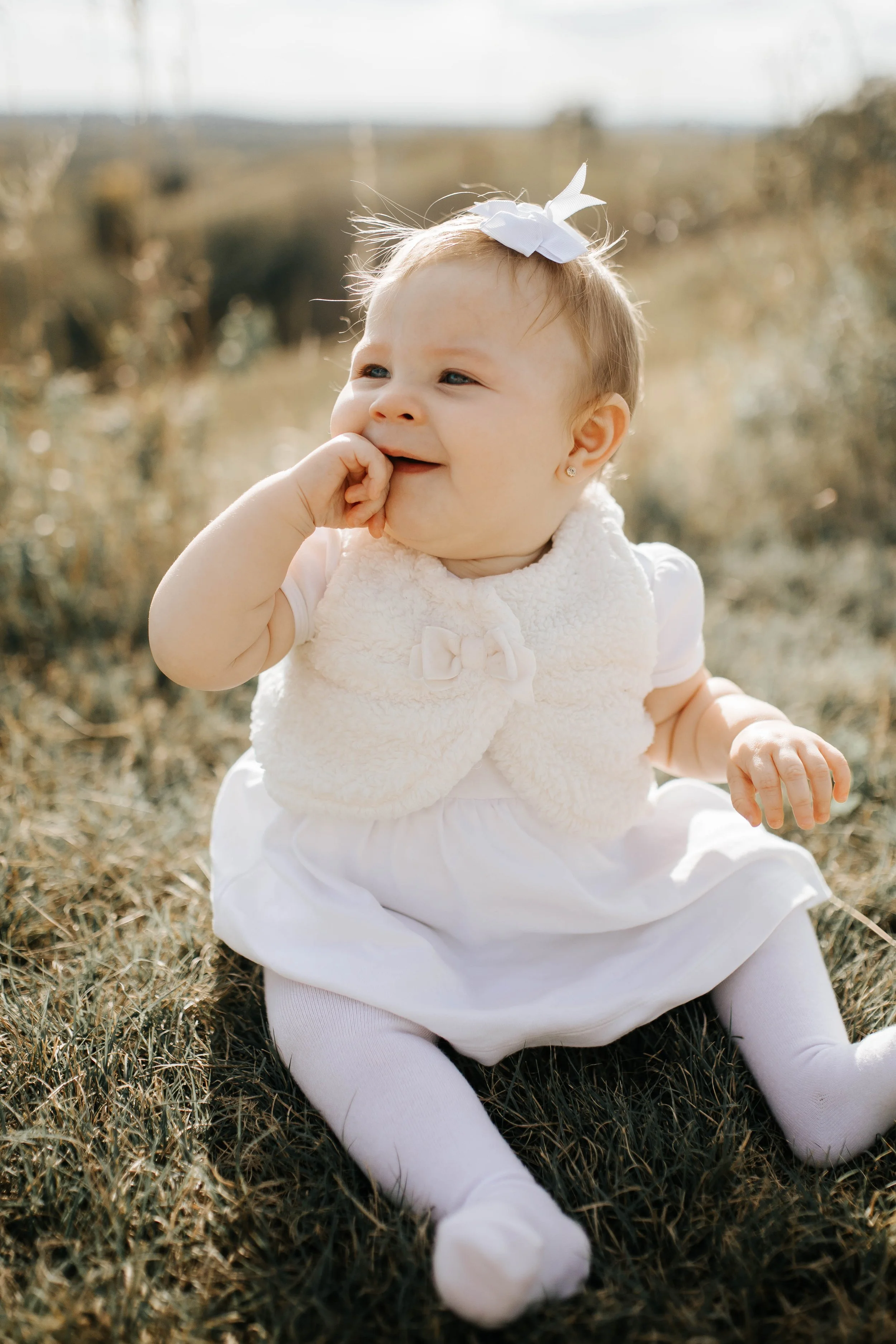 A young girl sitting on grass outdoors with a white bow on her head, wearing a white dress, a fluffy cream vest, and white tights, smiling and touching her mouth with her hand.