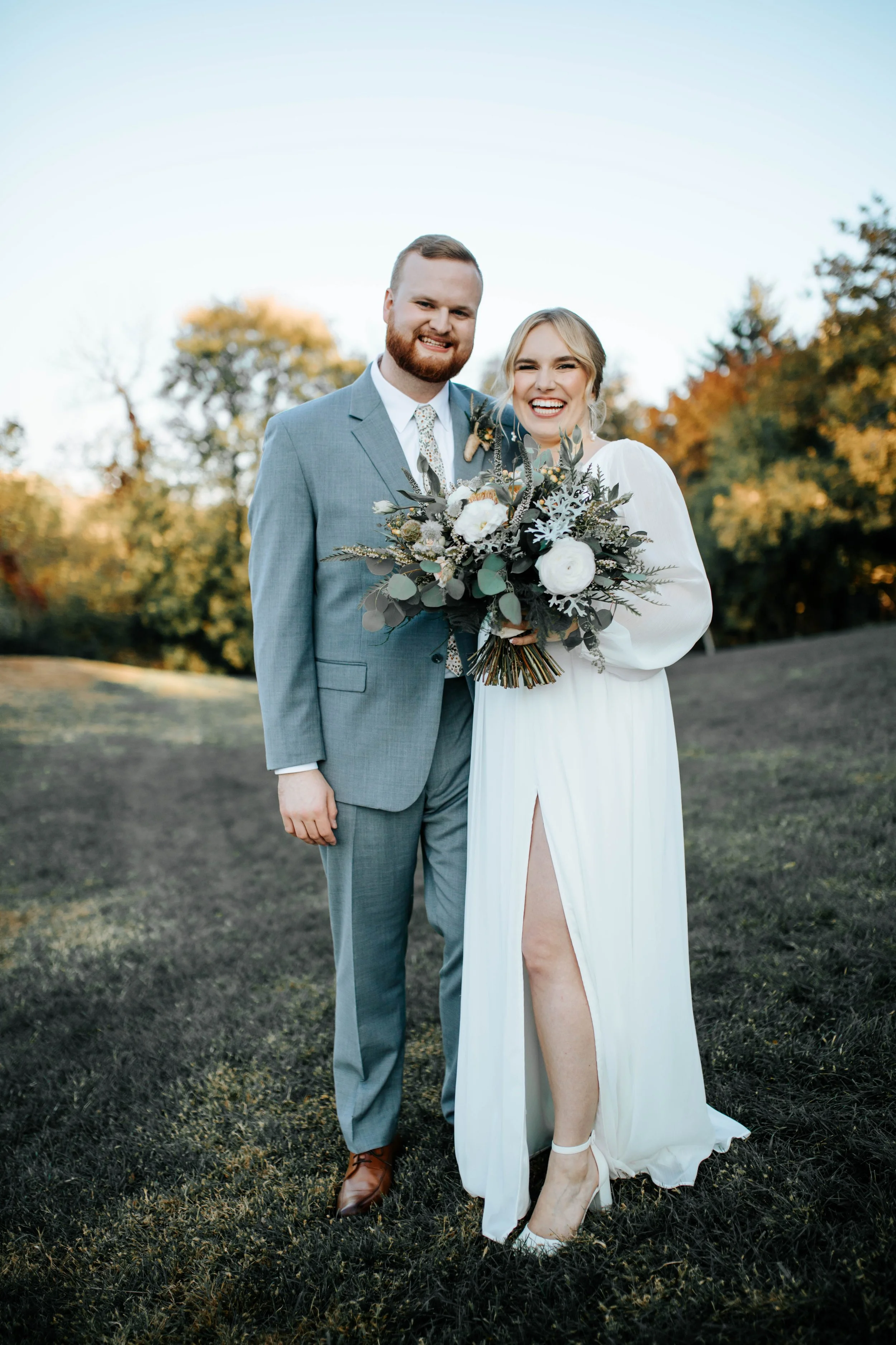 A smiling bride and groom standing outdoors with trees in the background, the bride holding a bouquet of flowers, dressed in a white wedding gown, and the groom in a light gray suit.