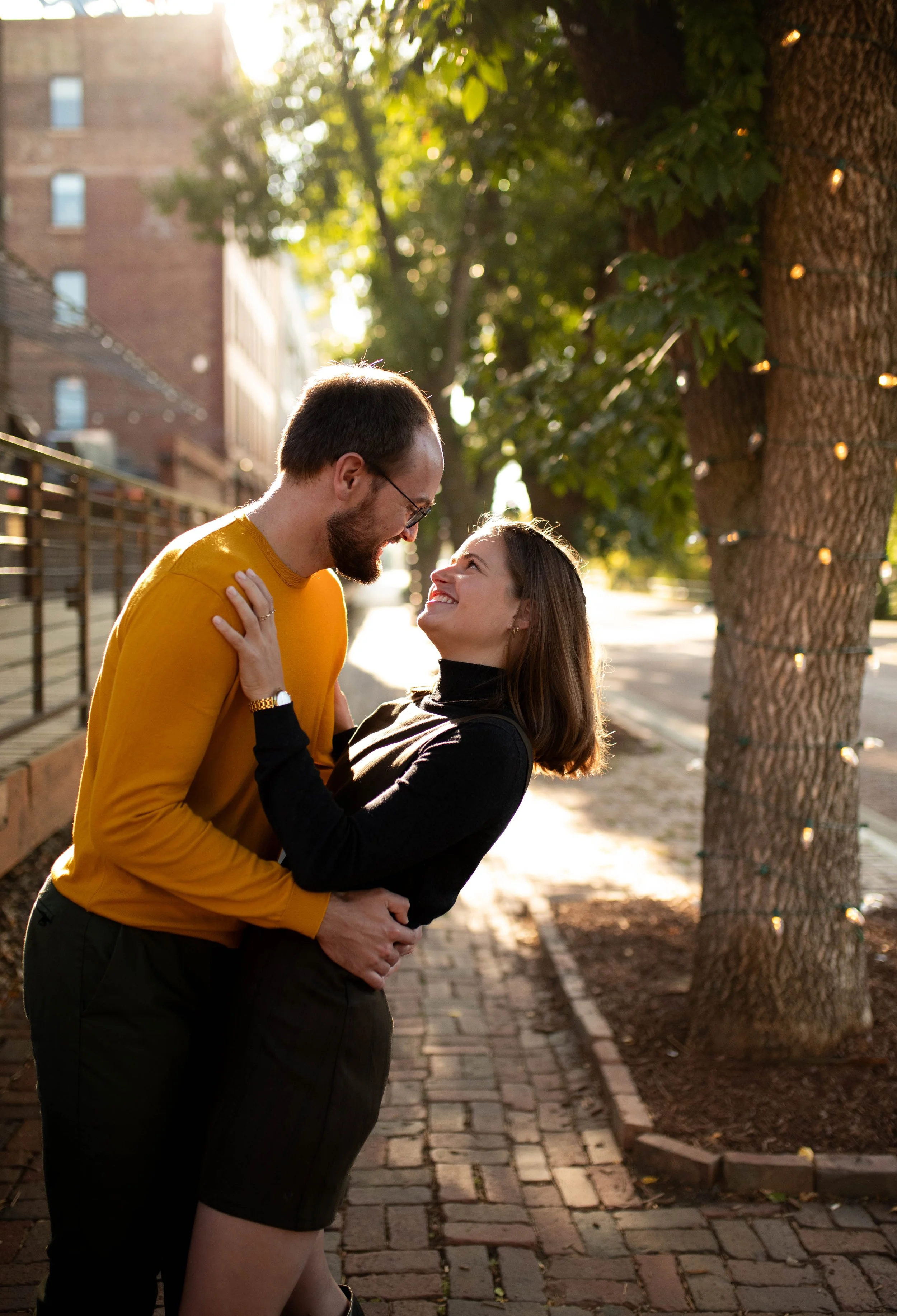 A couple smiling and embracing outdoors on a brick sidewalk, with a large tree decorated with string lights in the background.