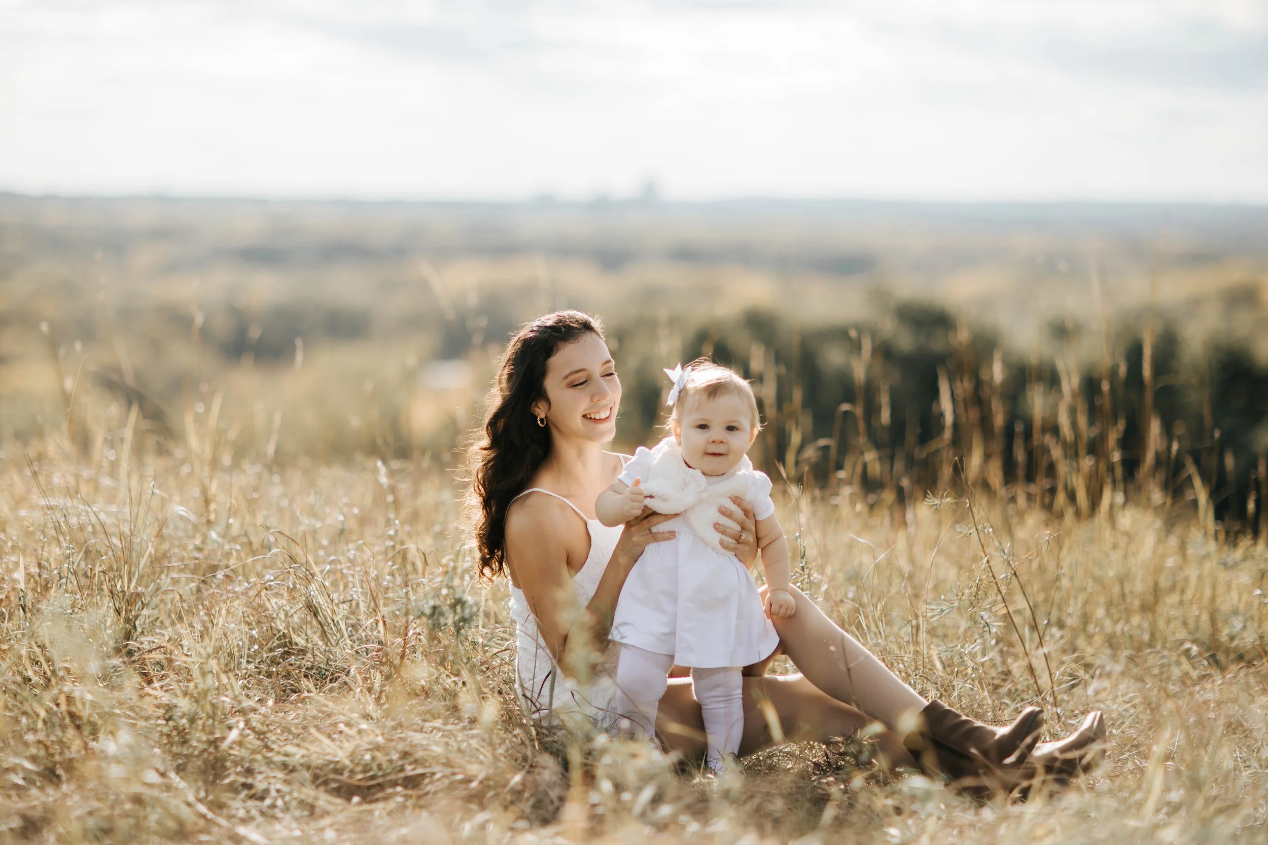 Young woman and a baby girl sitting in a grassy field on a sunny day, smiling and enjoying the outdoors.