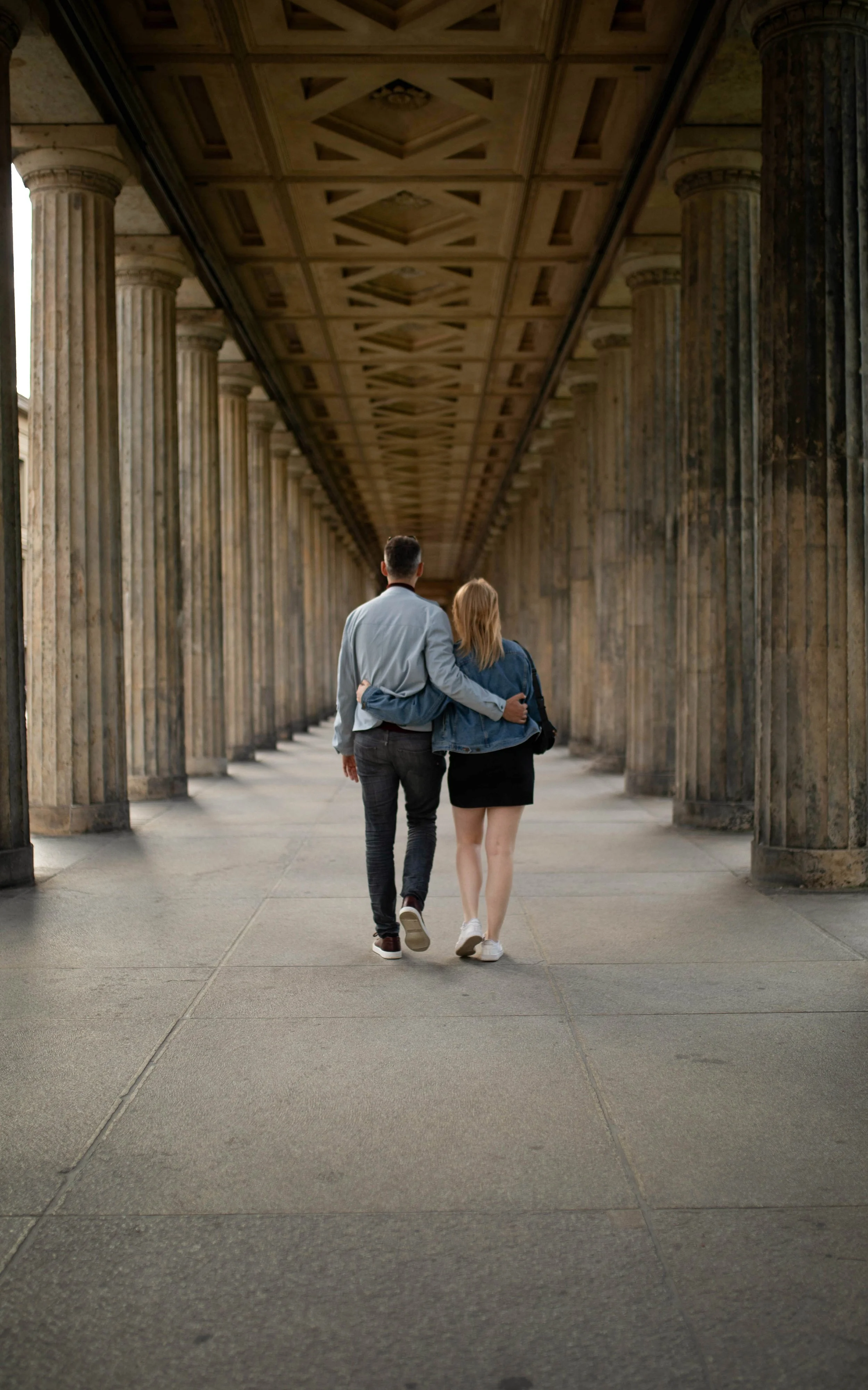 A couple walking arm in arm through a classical building with tall columns and detailed ceiling.