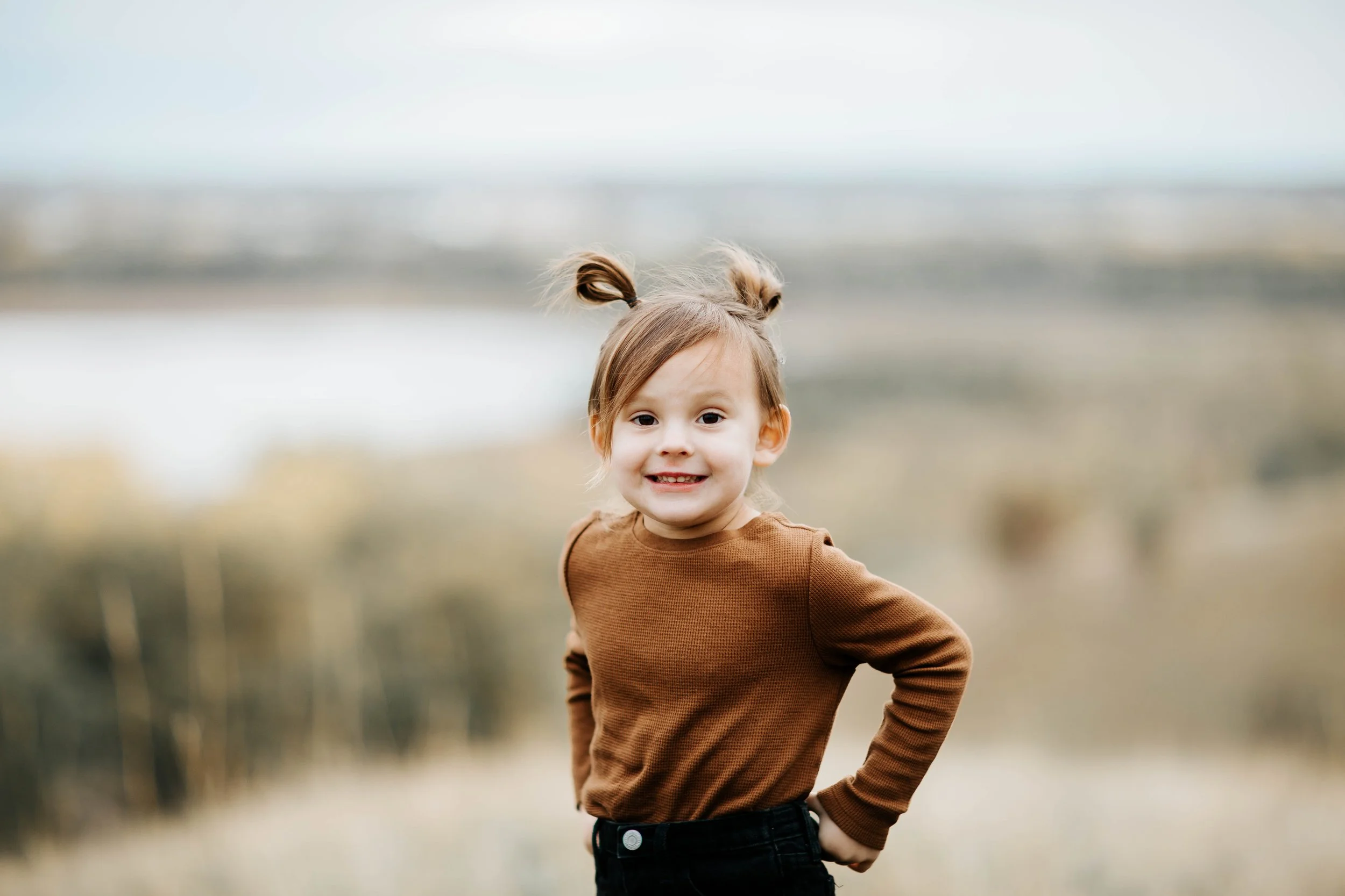 A young girl with light brown hair styled in two messy buns, wearing a brown long-sleeve shirt and black pants, smiling and posing outdoors in front of a blurred natural landscape.