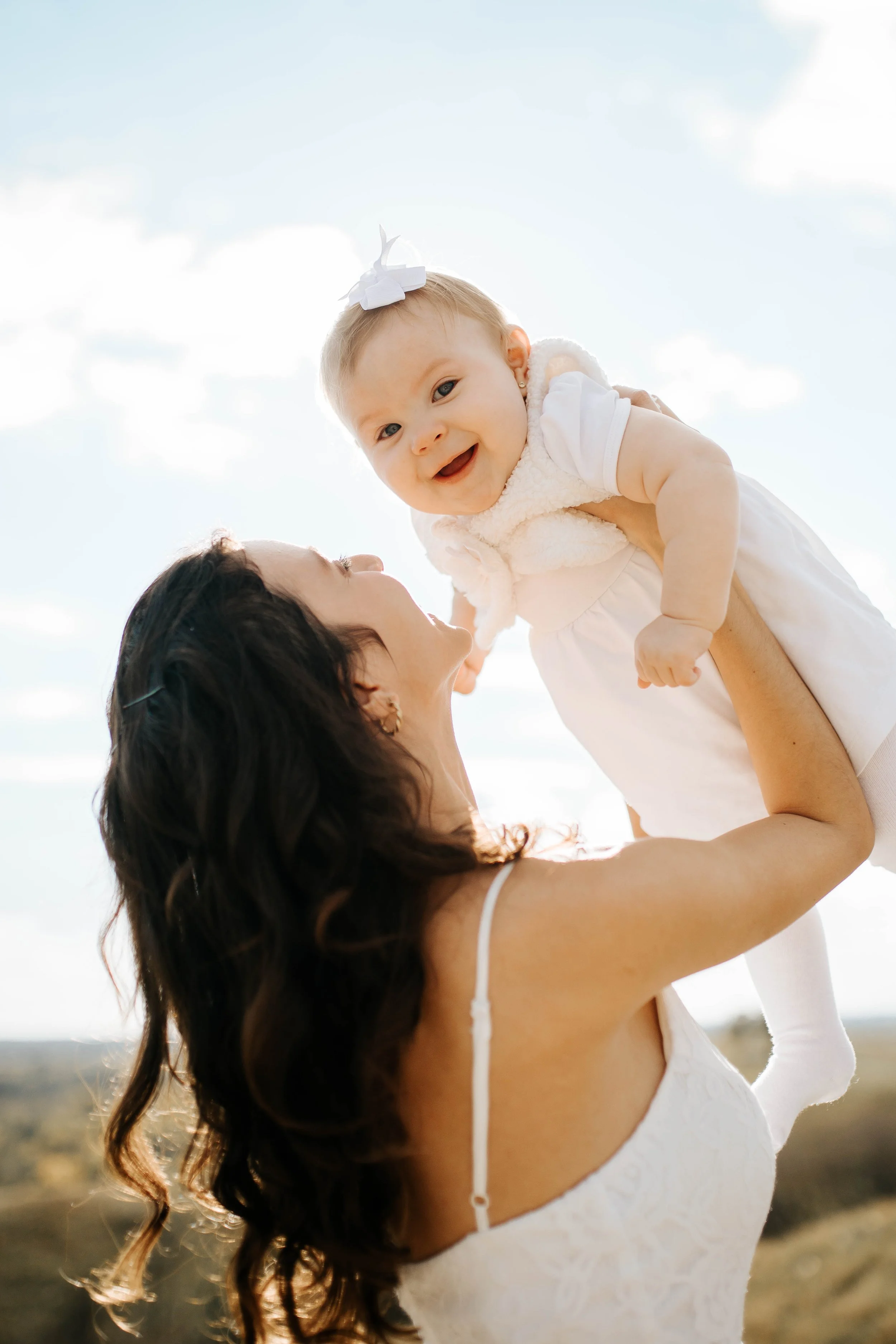A woman lifting a smiling baby girl outdoors during daytime. The baby is wearing a white dress and a bow headband, and the woman is dressed in a white lace top. The background shows a sky with scattered clouds.