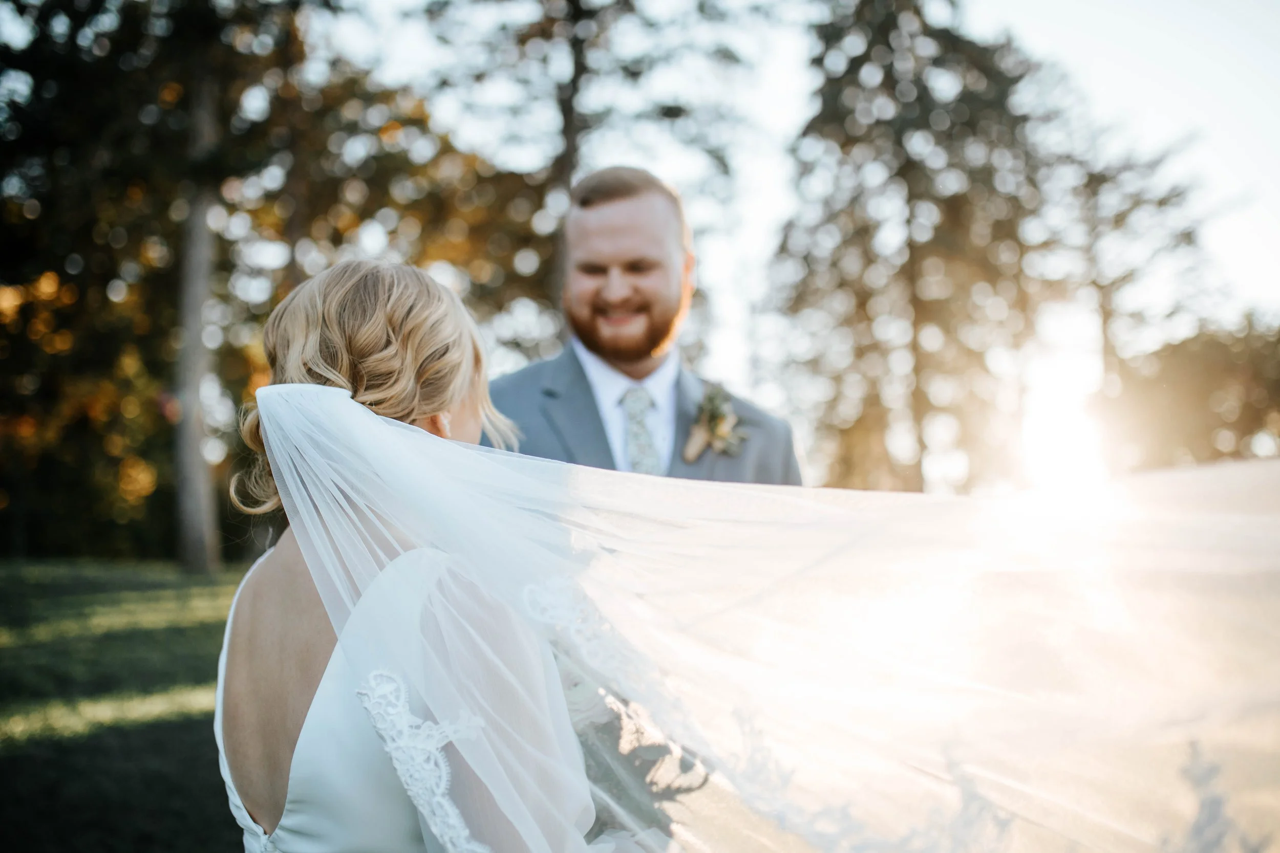 A bride with blonde hair in a wedding dress holding her veil, standing outdoors in a park with trees, as a smiling groom in a gray suit and tie looks at her during sunset.