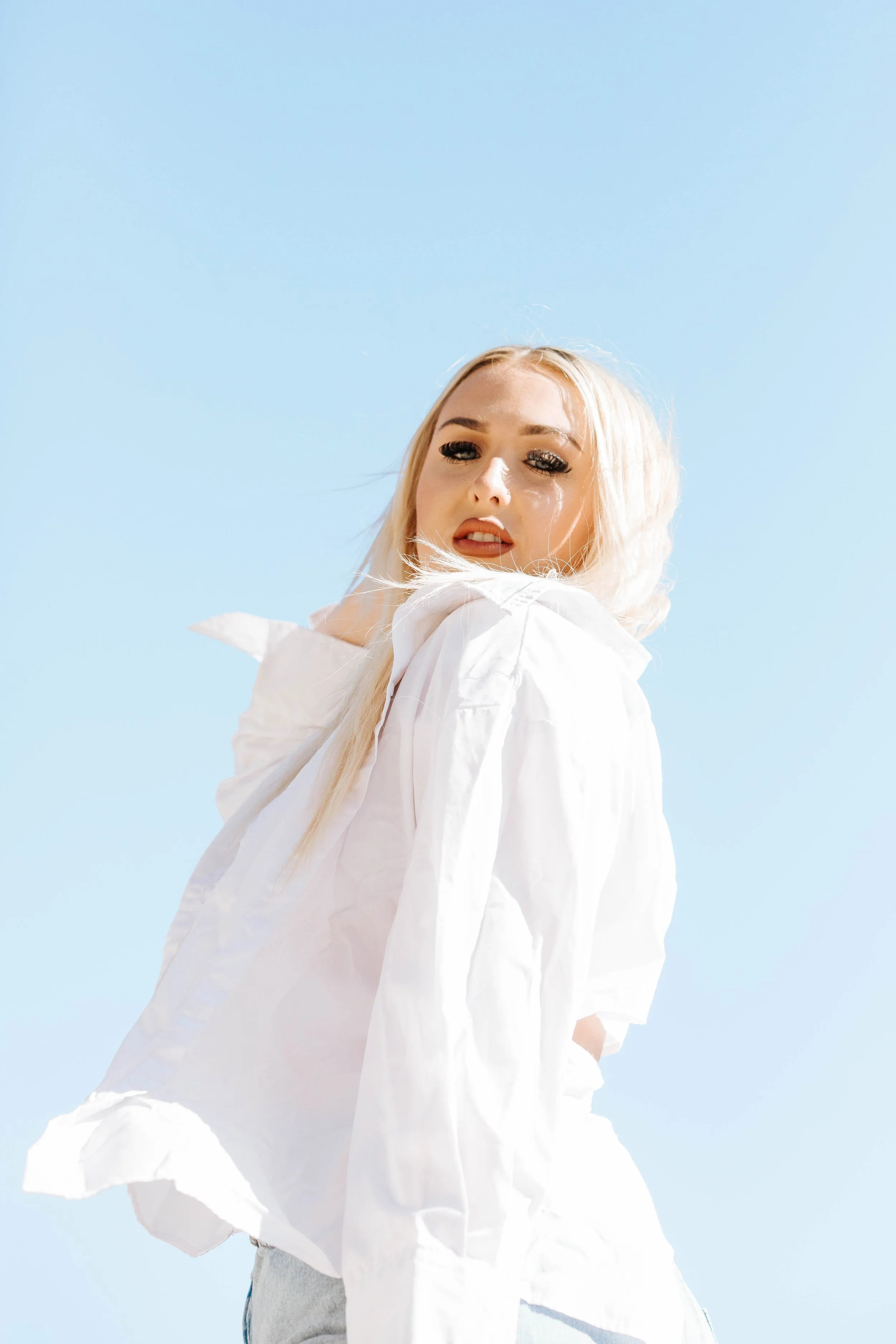A young woman with blonde hair wearing a white shirt against a clear blue sky