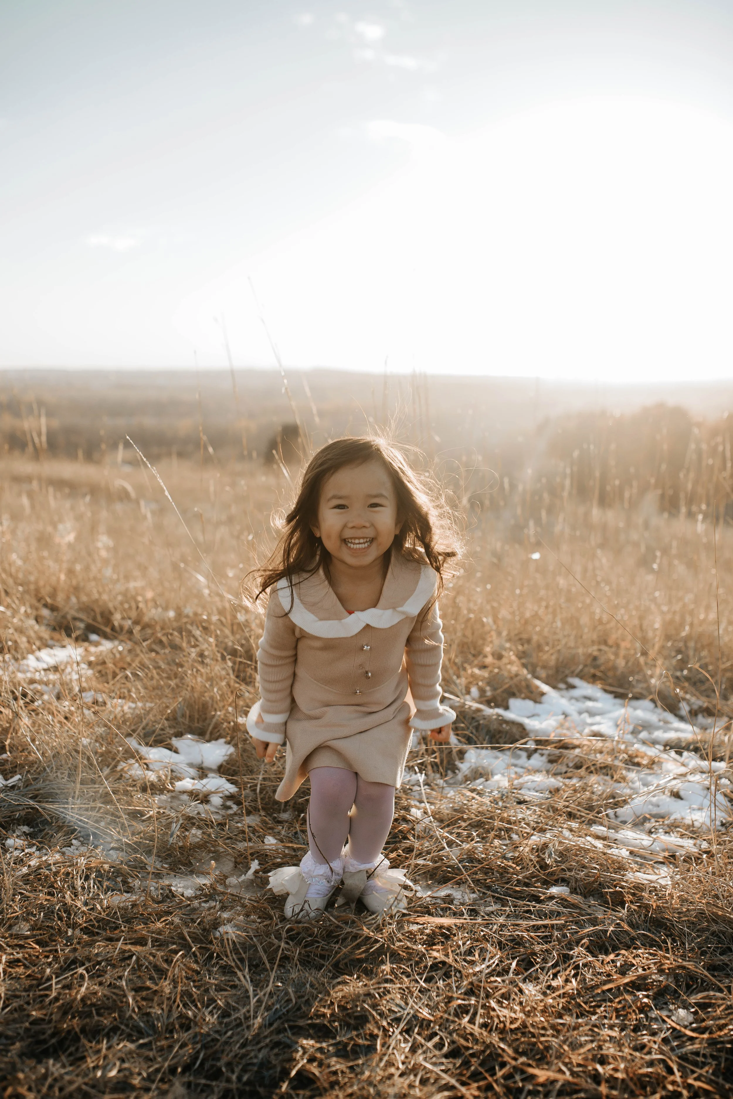 Young girl smiling and running in a field with tall, dry grass and patches of snow, during sunset.